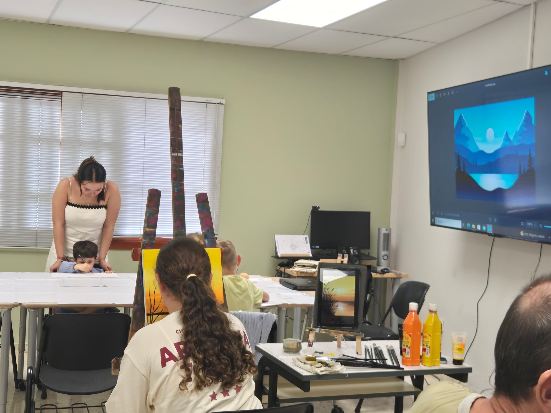 Personas en una clase de pintura; profesor y alumno en la mesa, pintando un paisaje mostrado en la pantalla, otros pintando en sus escritorios.