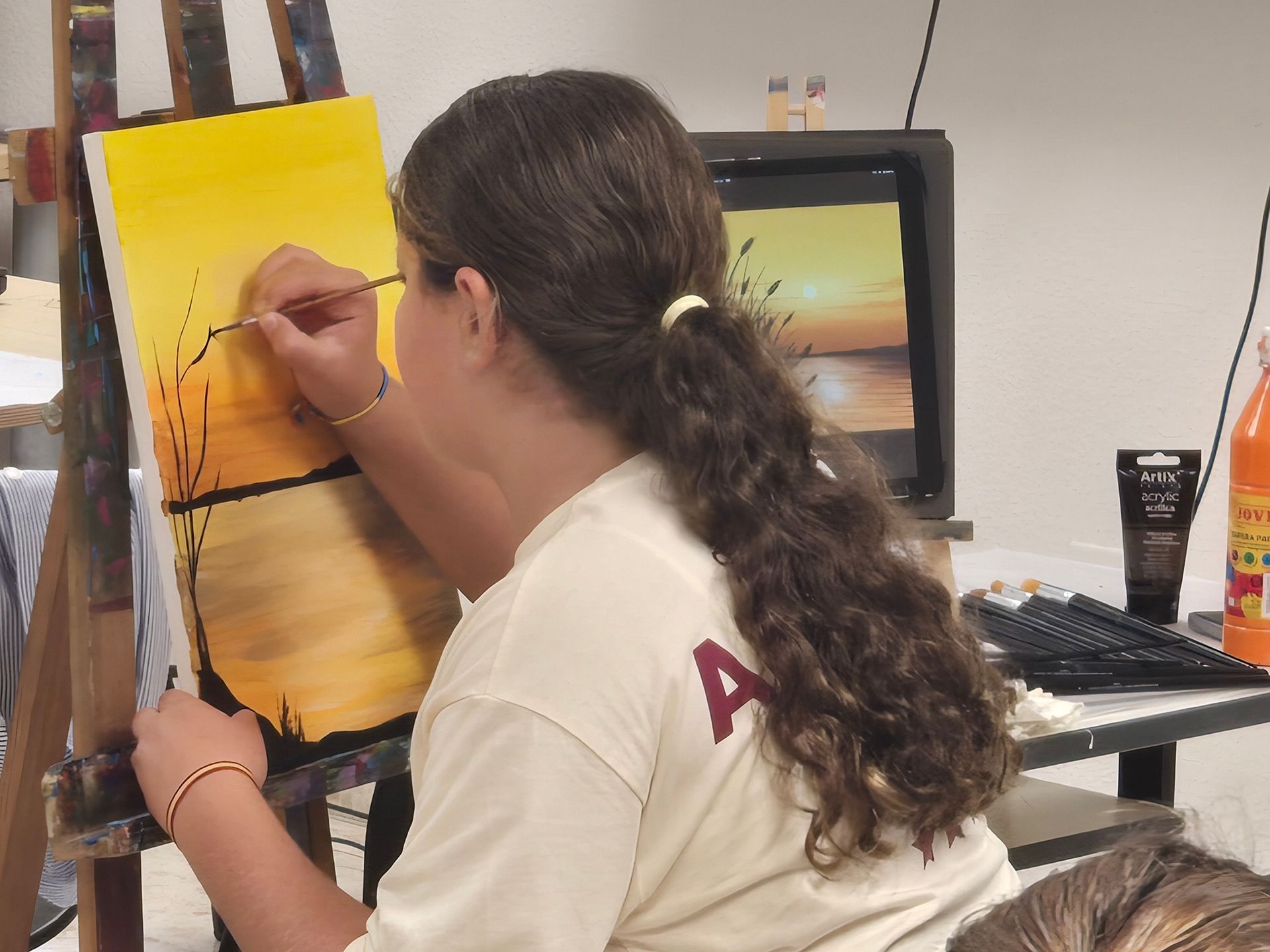 Una persona pinta un atardecer en un lienzo con un caballete. Lleva una camisa blanca y tiene el pelo castaño y rizado.