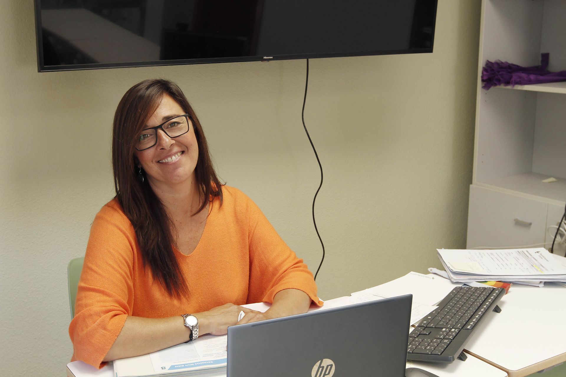 Una mujer con un suéter naranja sonríe en un escritorio con una computadora portátil.