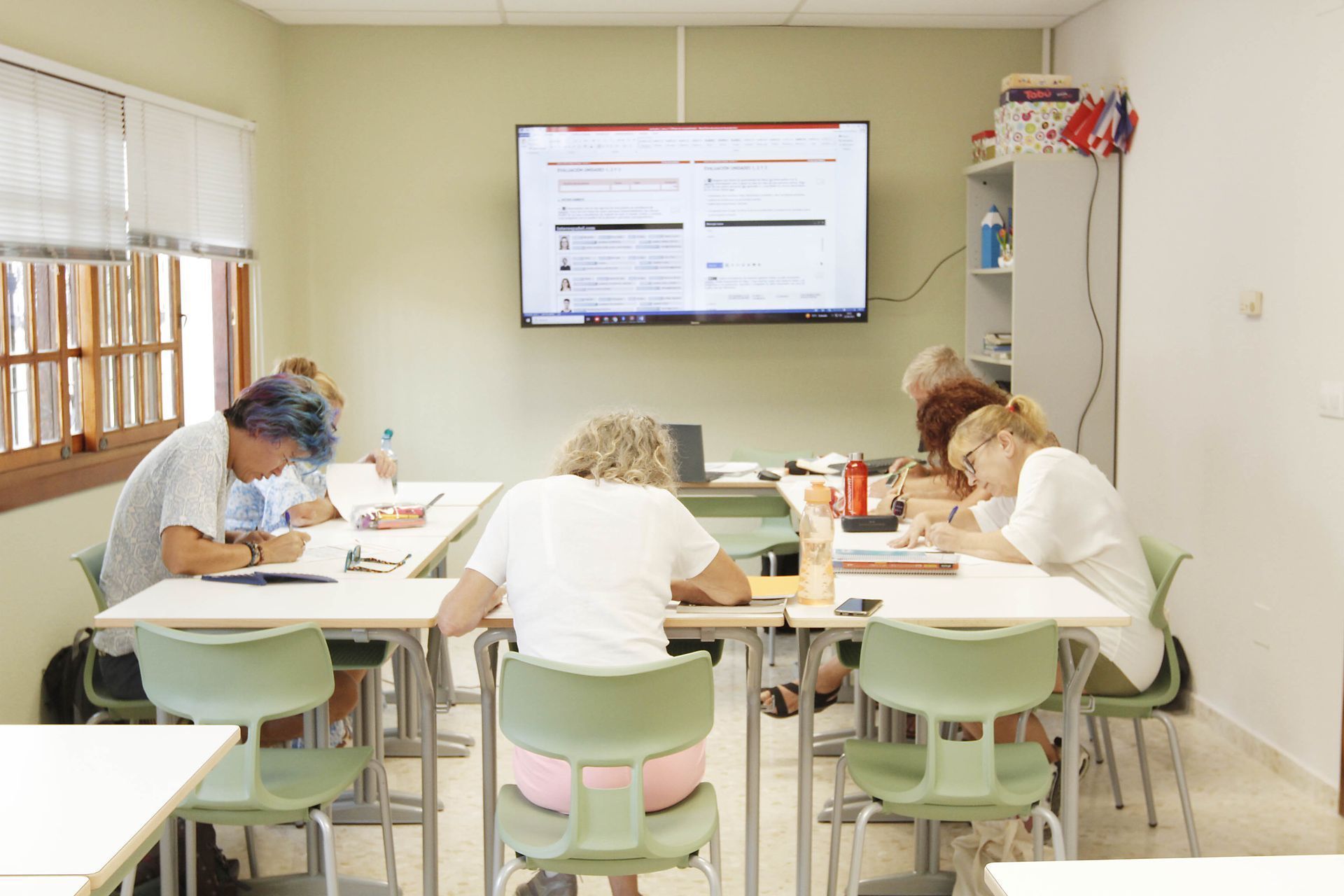 Personas escribiendo en escritorios en un aula, una pantalla muestra un sitio web.