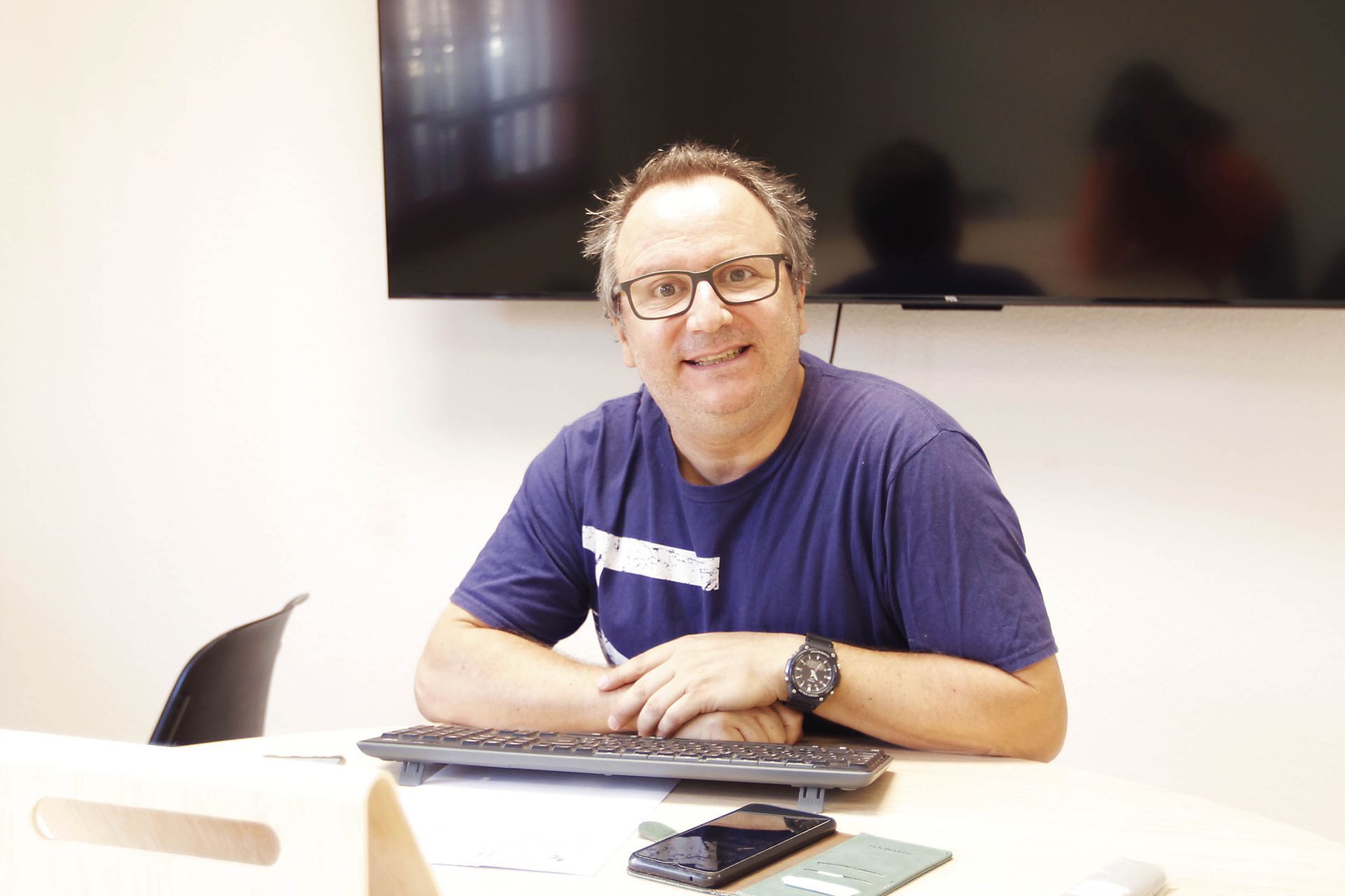 Hombre con gafas, sonriendo, sentado en un escritorio con un teclado, un teléfono y un reloj inteligente.