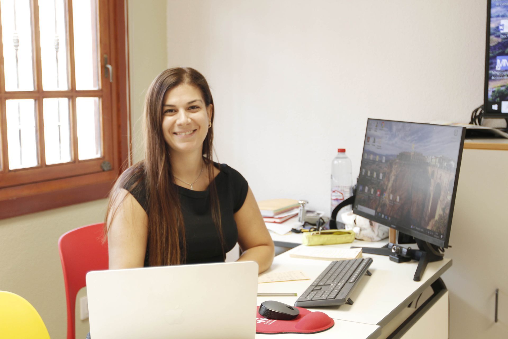 Mujer sonriendo en un escritorio con una computadora portátil y un monitor, en una oficina bien iluminada.