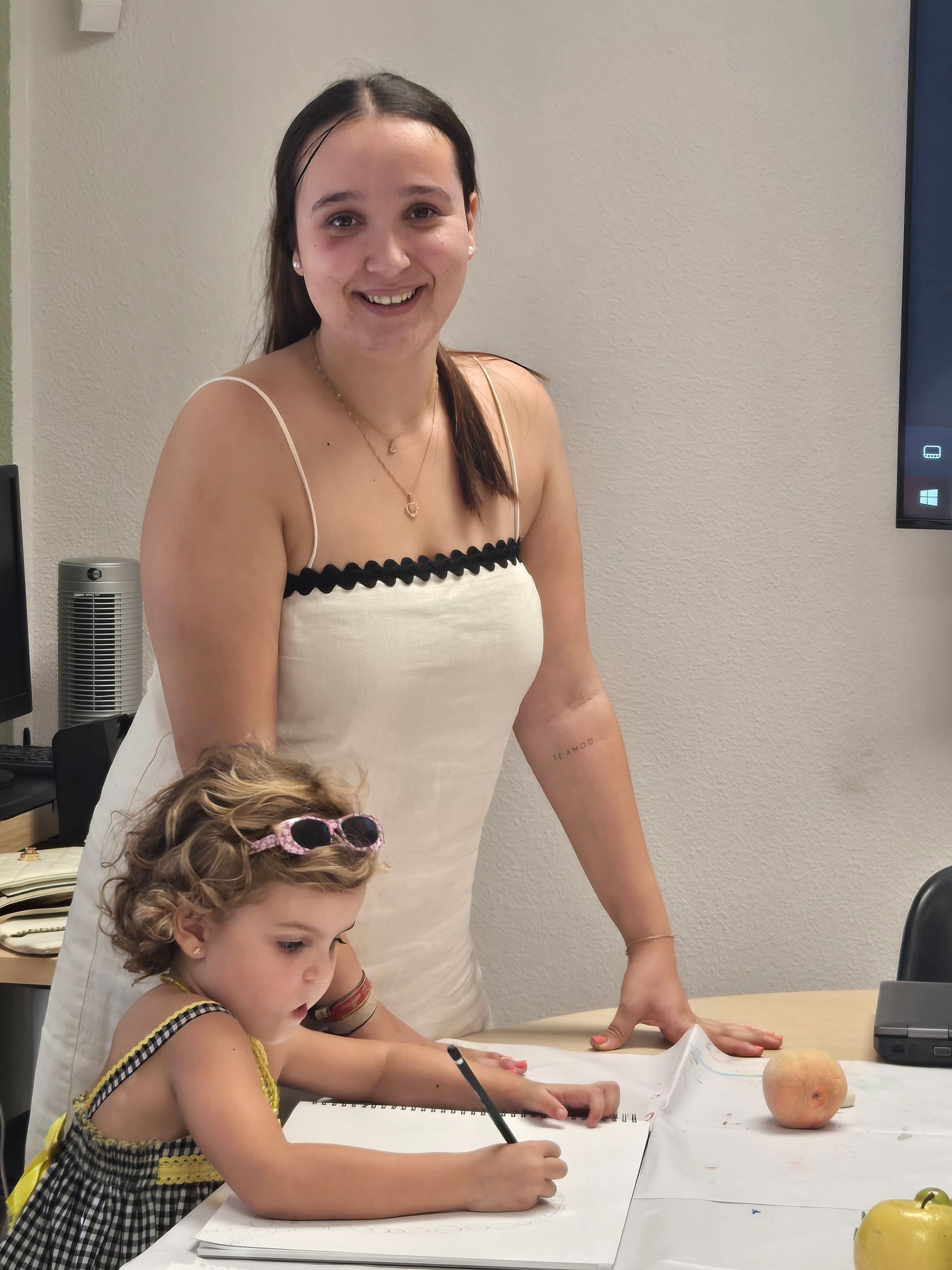 Una mujer sonríe mientras un niño dibuja en una mesa. Vestido blanco, niño con gafas de sol, pared beige.