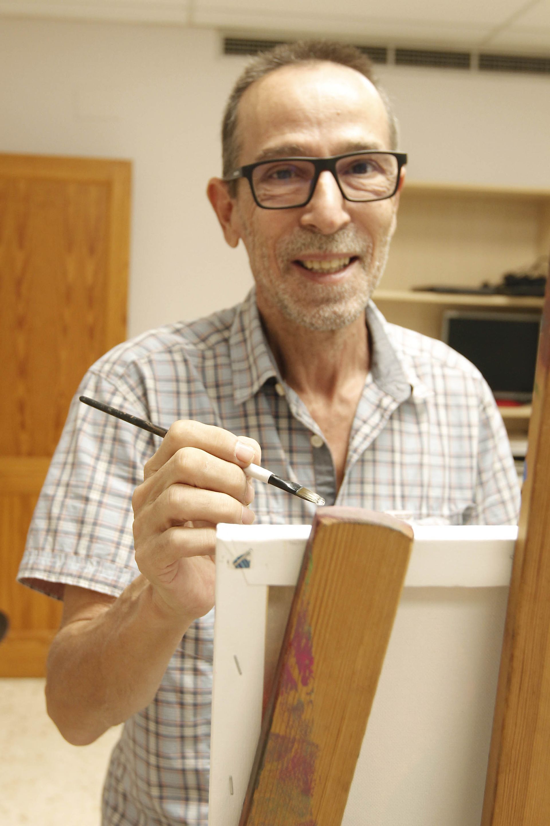 Hombre con gafas pintando sobre lienzo, sonriendo, en un estudio con caballete de madera, vistiendo una camisa a cuadros.