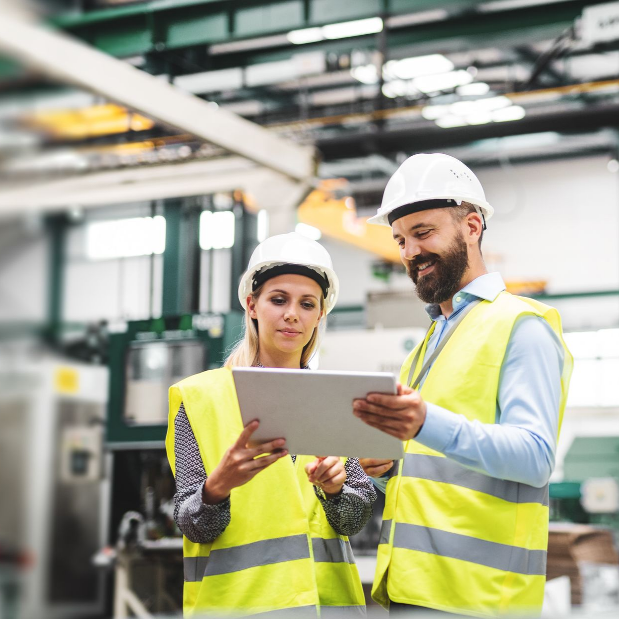 Deux personnes portant des casques et des gilets de sécurité regardent une tablette dans un environnement industriel.