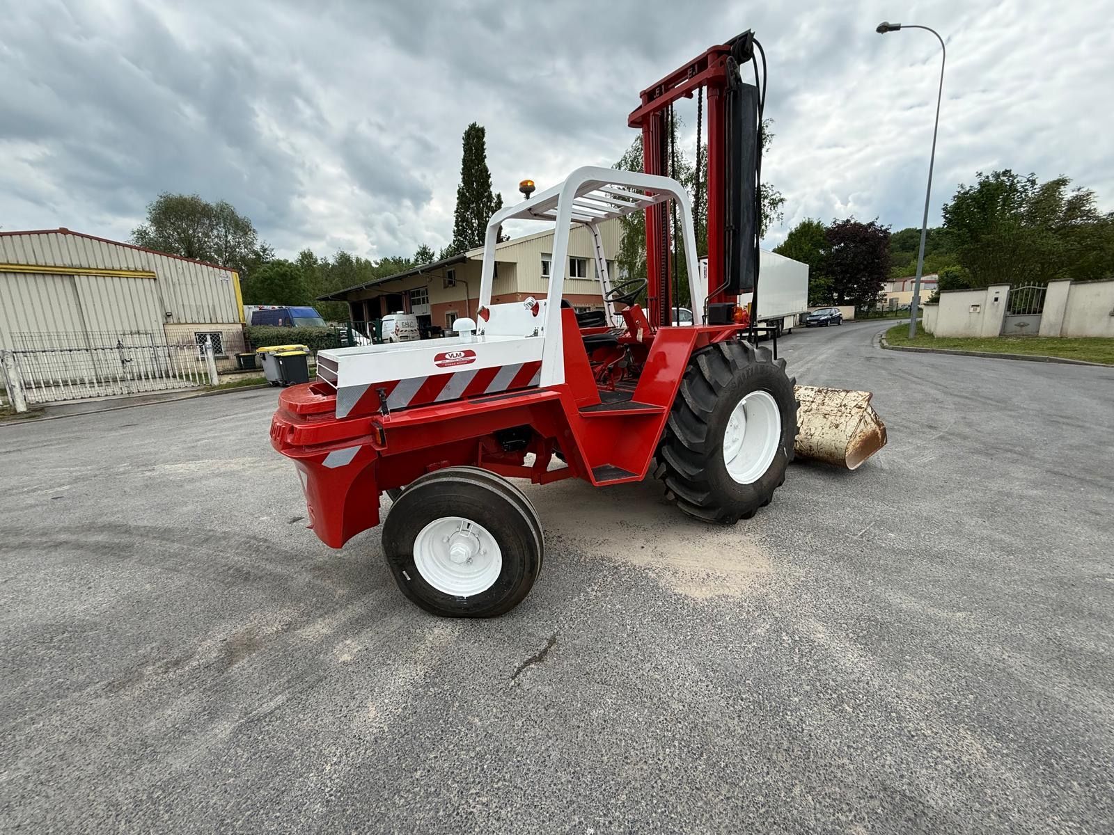 Un chariot élévateur rouge et blanc est stationné sur un terrain gravillonné près d'un bâtiment industriel.