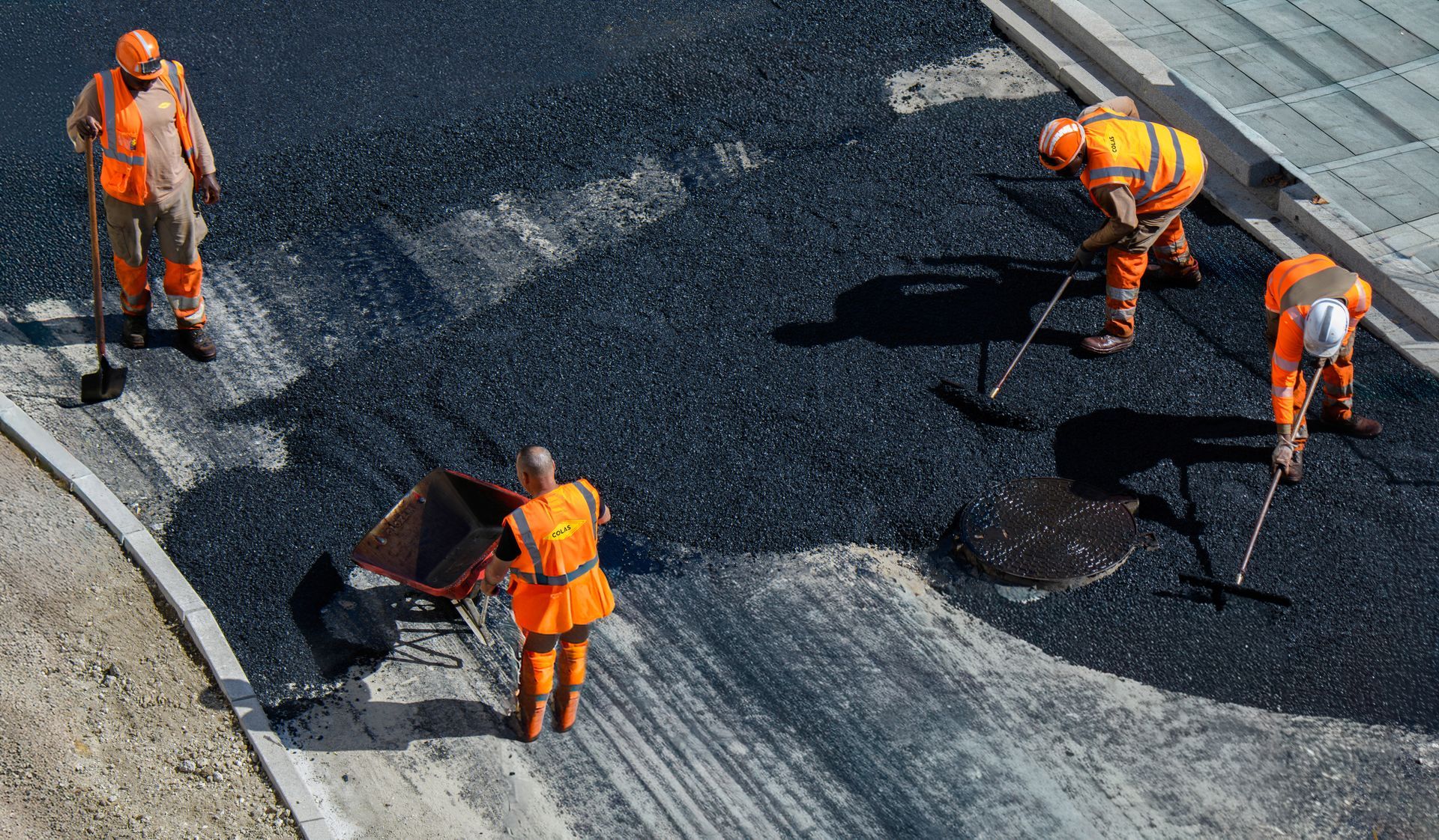 Des ouvriers en tenue de sécurité orange réparent l'asphalte d'une route en virage.