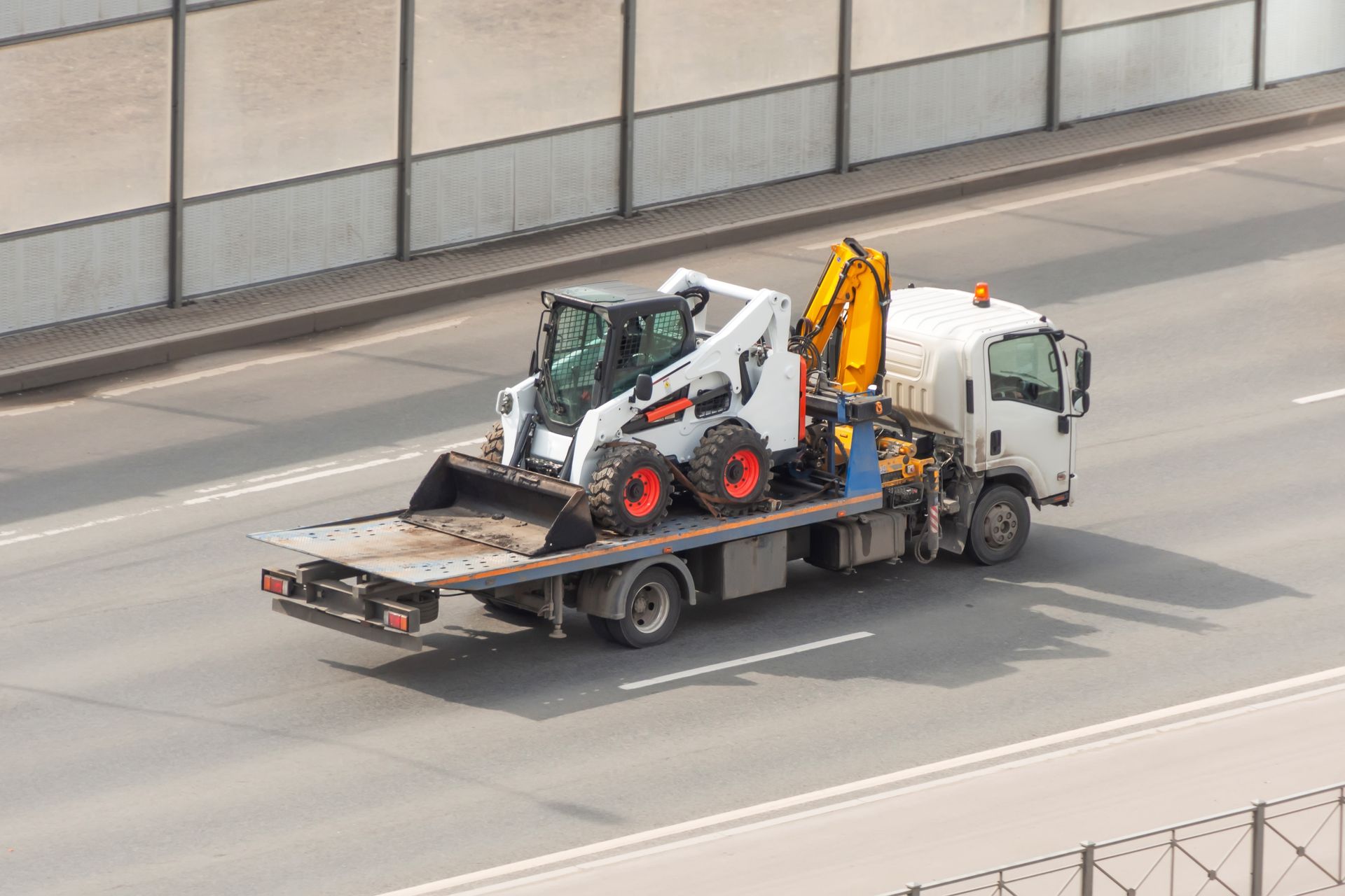 Camion de dépannage blanc à plateau transportant une chargeuse compacte et une petite pelle mécanique sur une autoroute