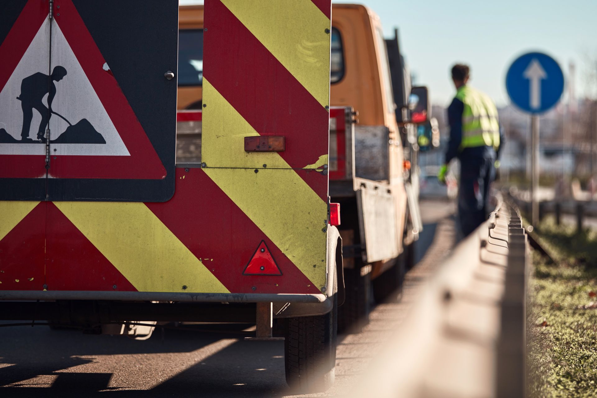 Panneau de travaux et barrière en bord de route, avec des ouvriers et une camionnette en arrière-plan.