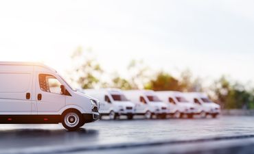 Des camionnettes de livraison blanches garées en rang à l'extérieur, sur une route ensoleillée.