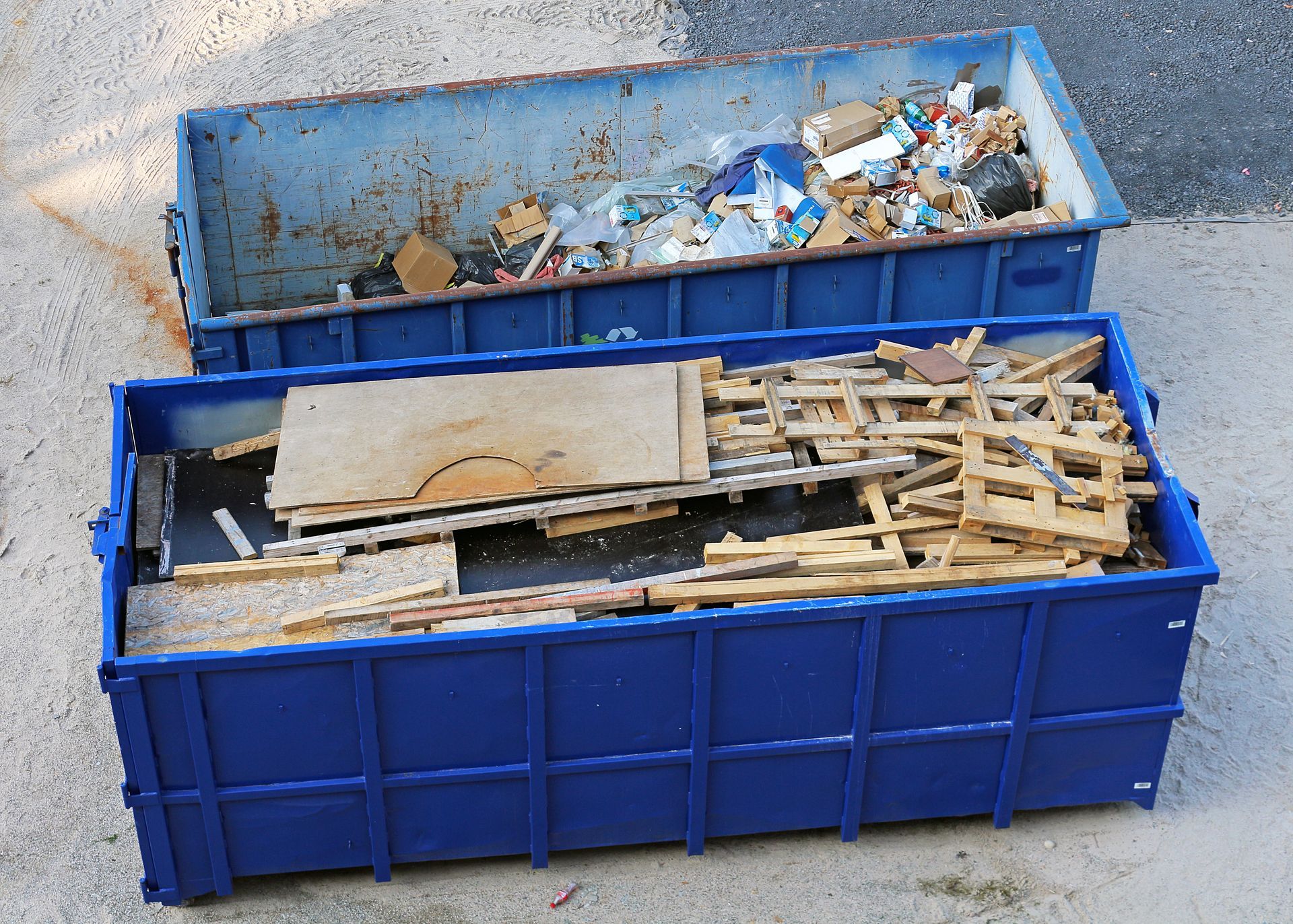Deux bennes bleues remplies de débris de construction et de chutes de bois à l'extérieur.