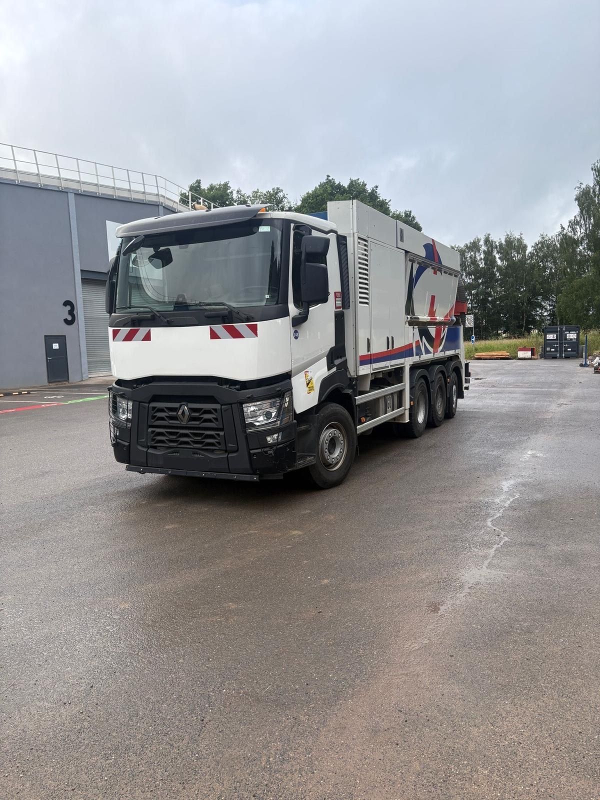 Camion-poubelle blanc stationné sur un terrain humide à côté d'un bâtiment industriel gris.