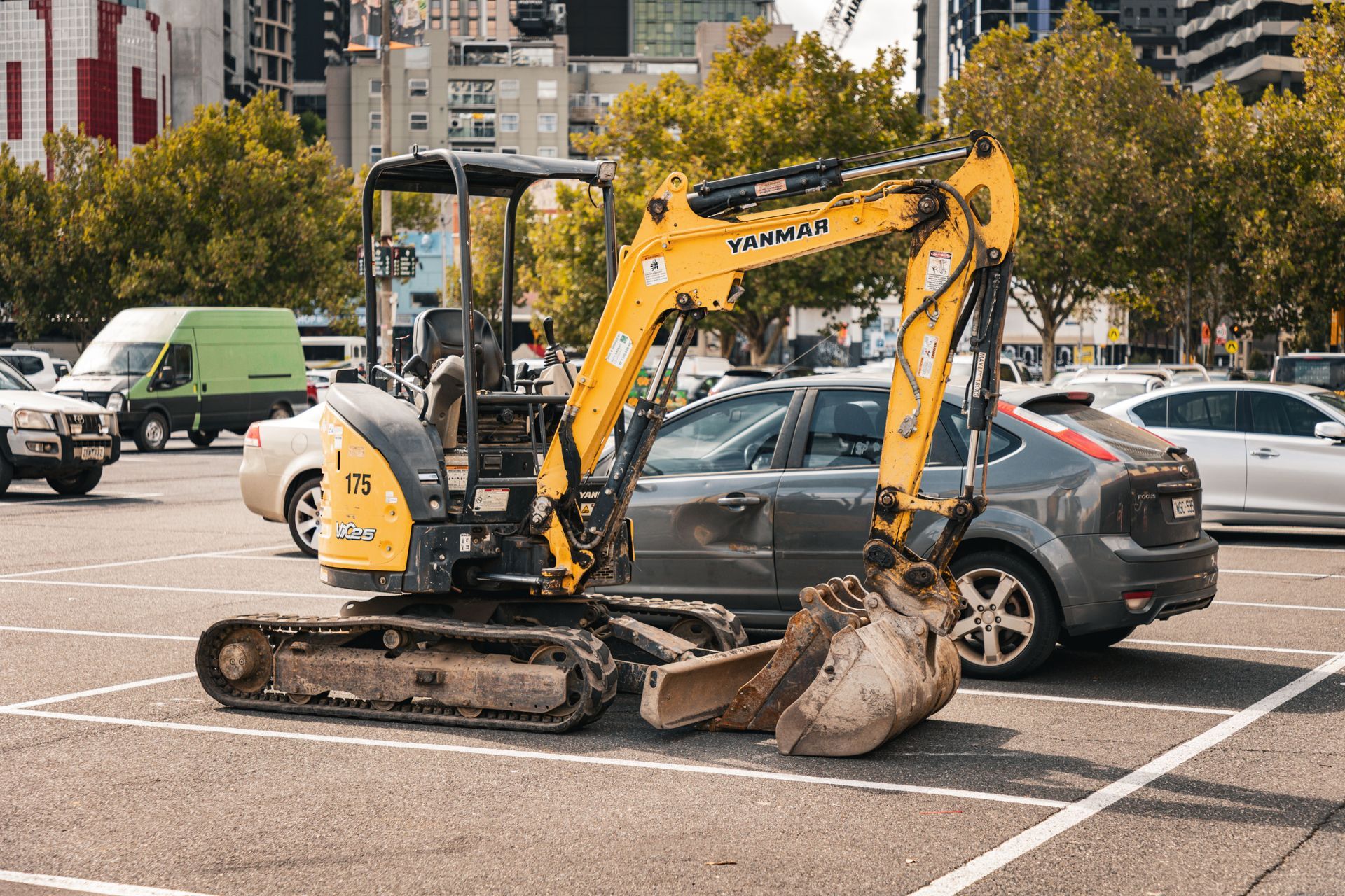Une minipelle jaune est garée sur un terrain municipal à côté d'une voiture grise et d'une camionnette verte, avec des bâtiments et des arbres en arrière-plan.