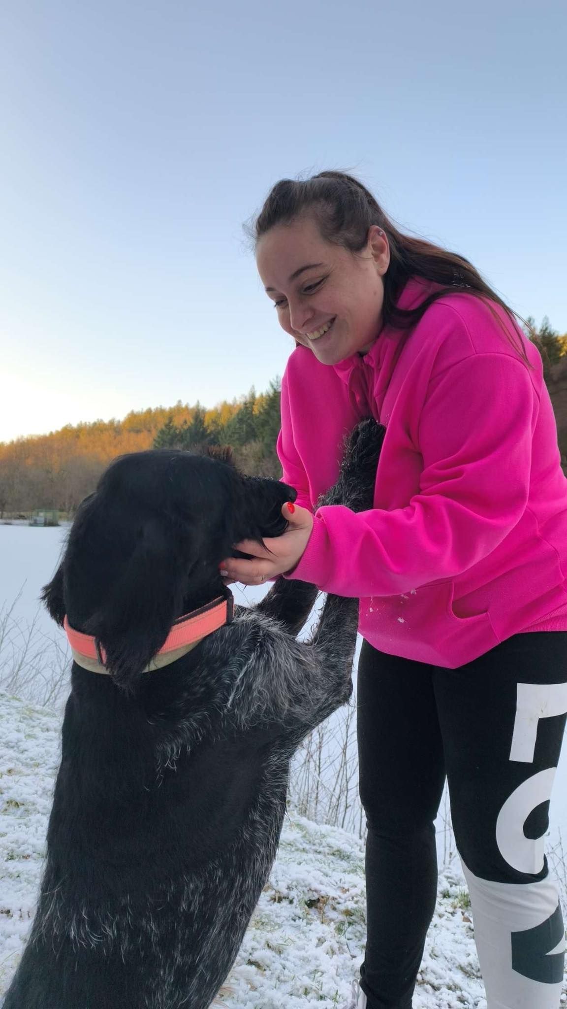 Une femme en sweat-shirt rose sourit en caressant un chien noir debout sur ses pattes arrière dans un champ enneigé.