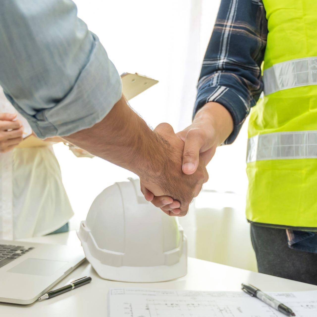 Un ouvrier serre la main à un client au-dessus d'un bureau où se trouvent un casque blanc et un ordinateur portable, ce qui laisse supposer la conclusion d'un accord de construction.