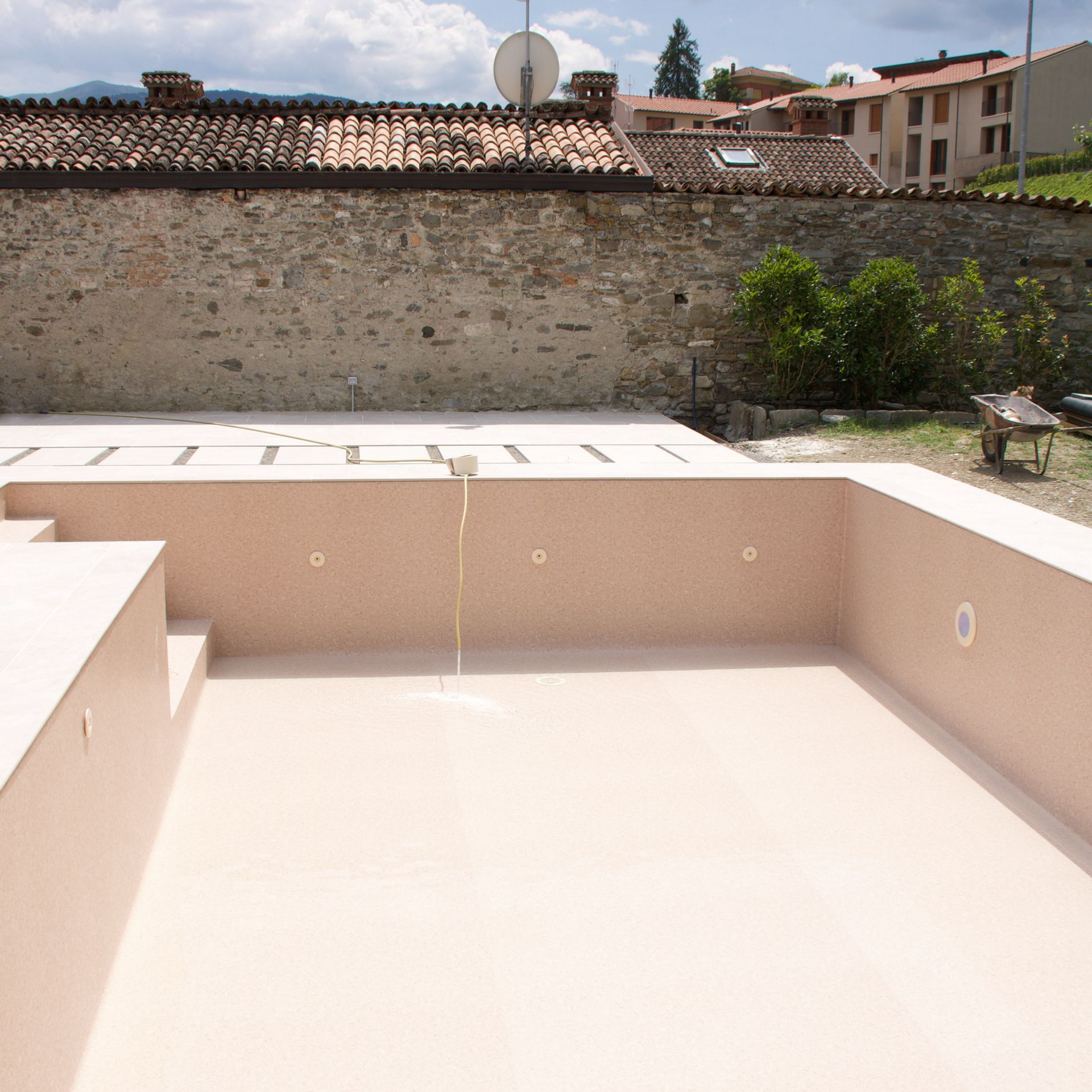 Piscine beige vide sous un soleil éclatant, avec un mur de pierre et des toits en arrière-plan.