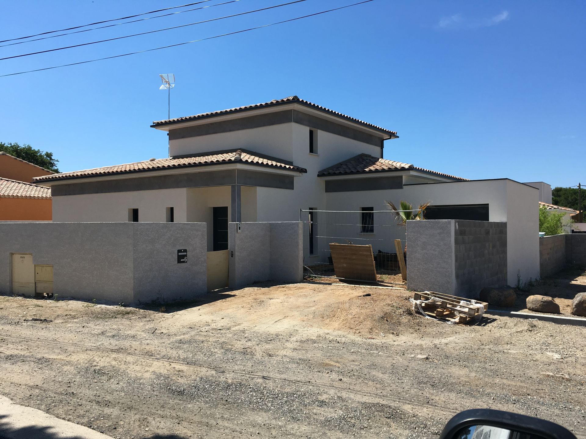Maison moderne en construction derrière un muret, au bord d'une route poussiéreuse, sous un ciel d'un bleu limpide.