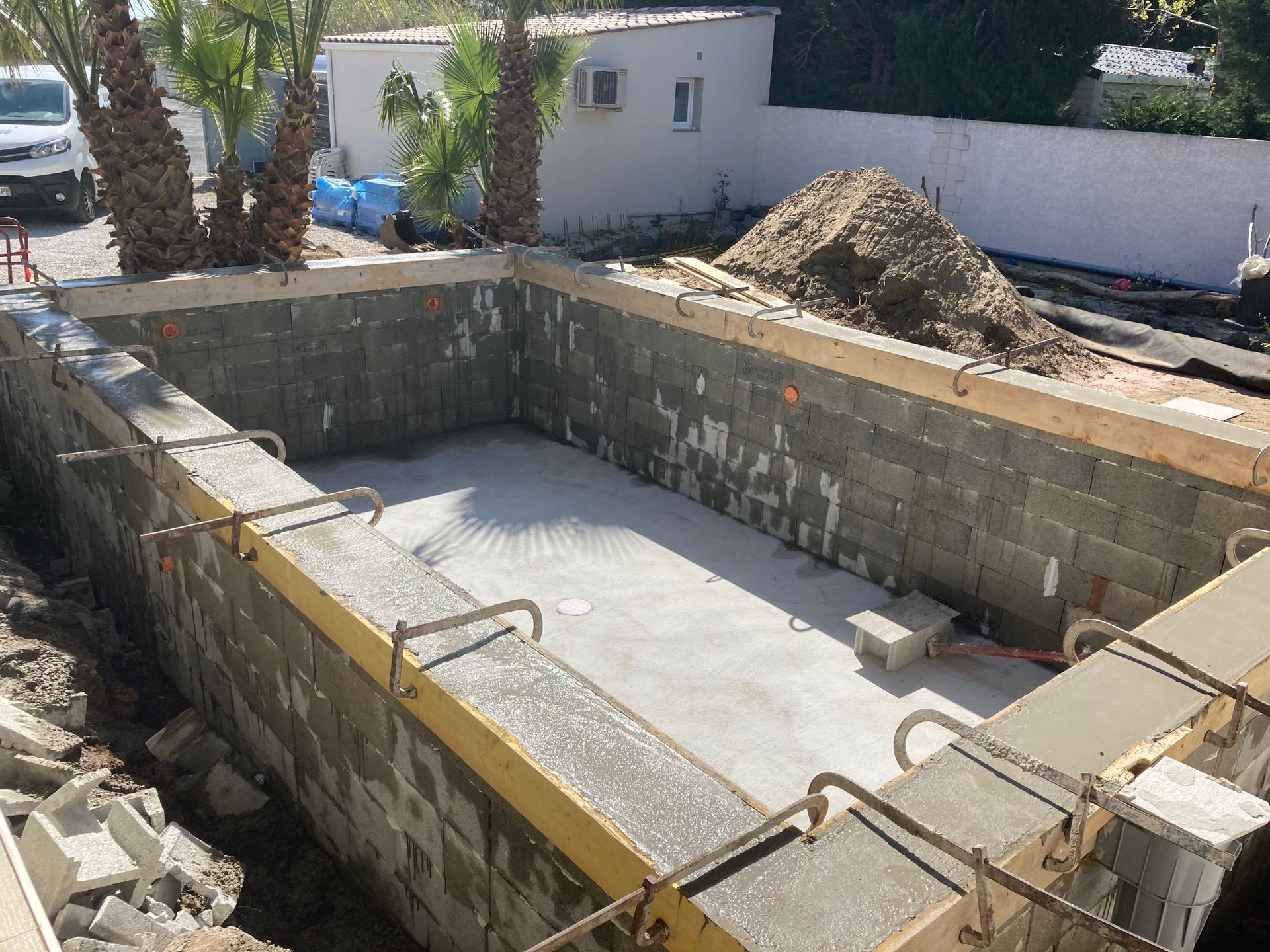 Piscine en béton en construction dans un jardin, avec des murs en parpaings et un tas de terre à proximité