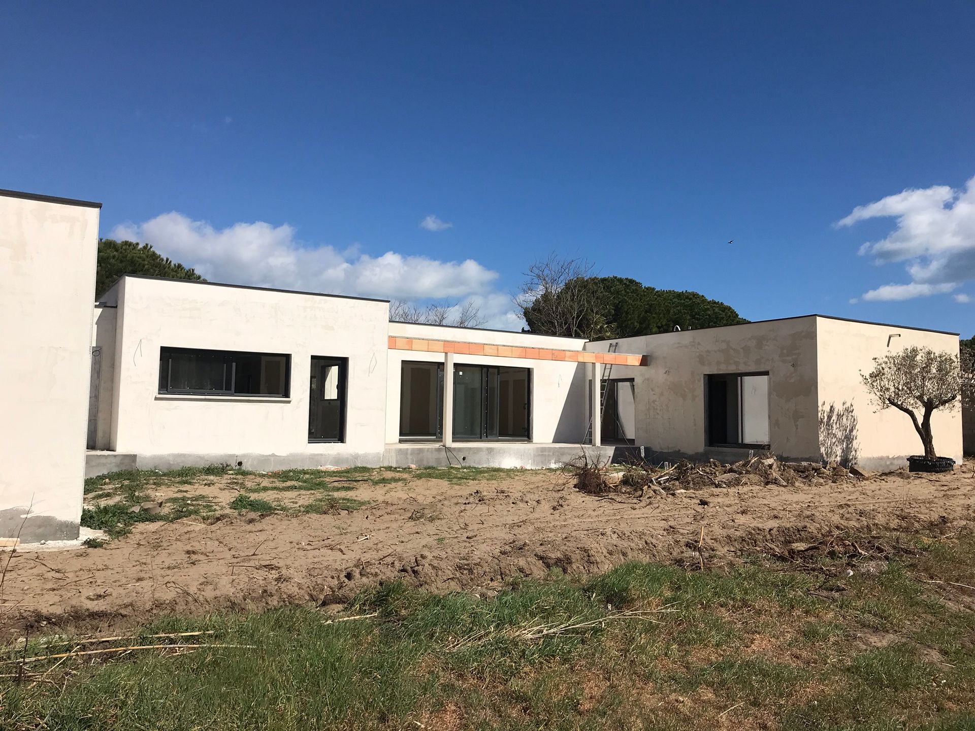 Maison moderne blanche avec toit plat et grandes fenêtres sur un terrain nu sous un ciel bleu.