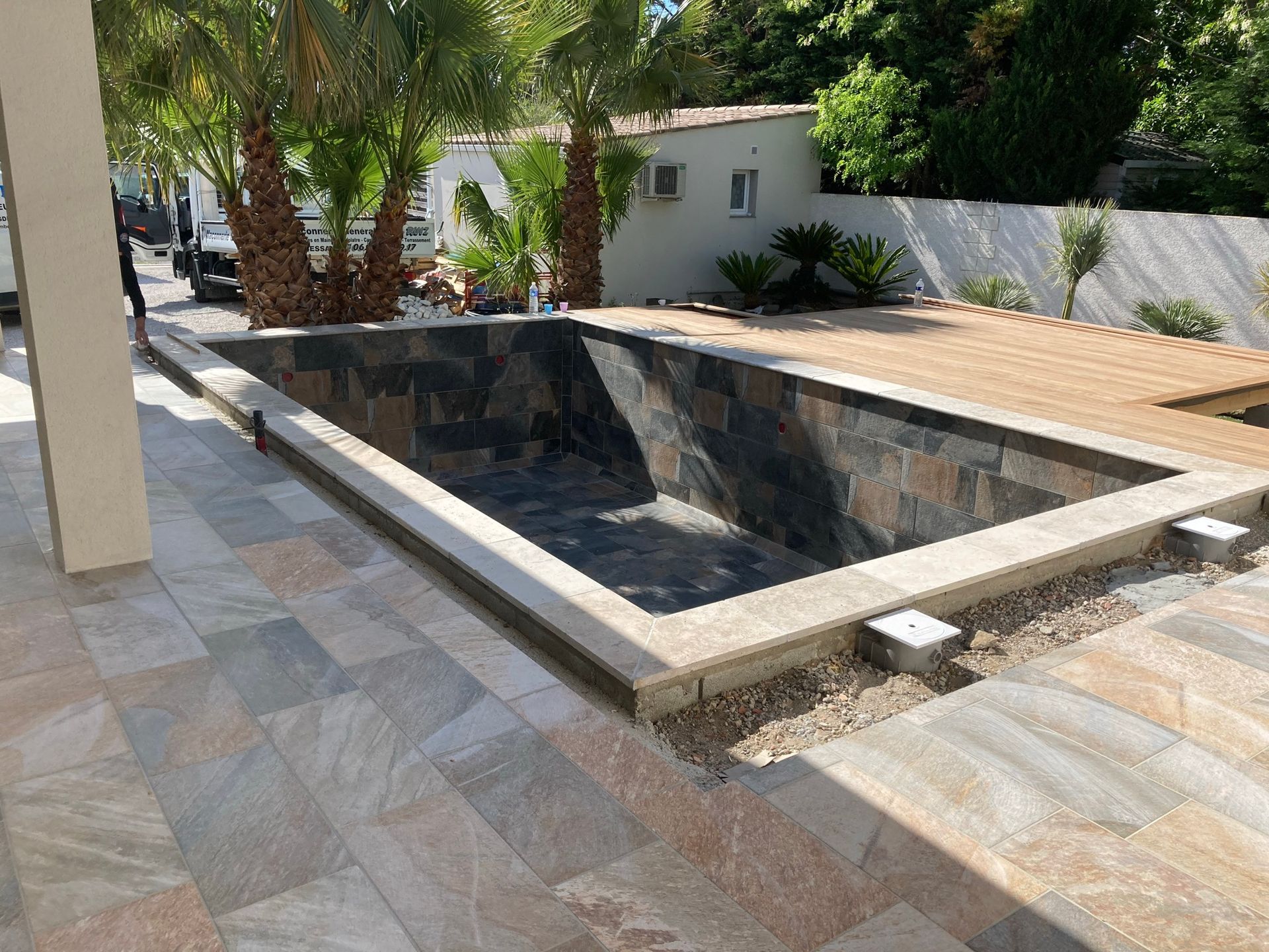 Piscine rectangulaire vide jouxtant une terrasse carrelée, avec arbres, terrasse en bois et mur blanc dans une cour.