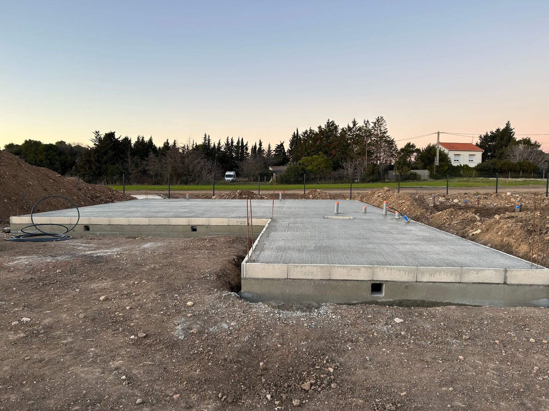 Dalles de fondations en béton sur un chantier de construction au crépuscule, avec un sol en terre battue et des arbres en arrière-plan.