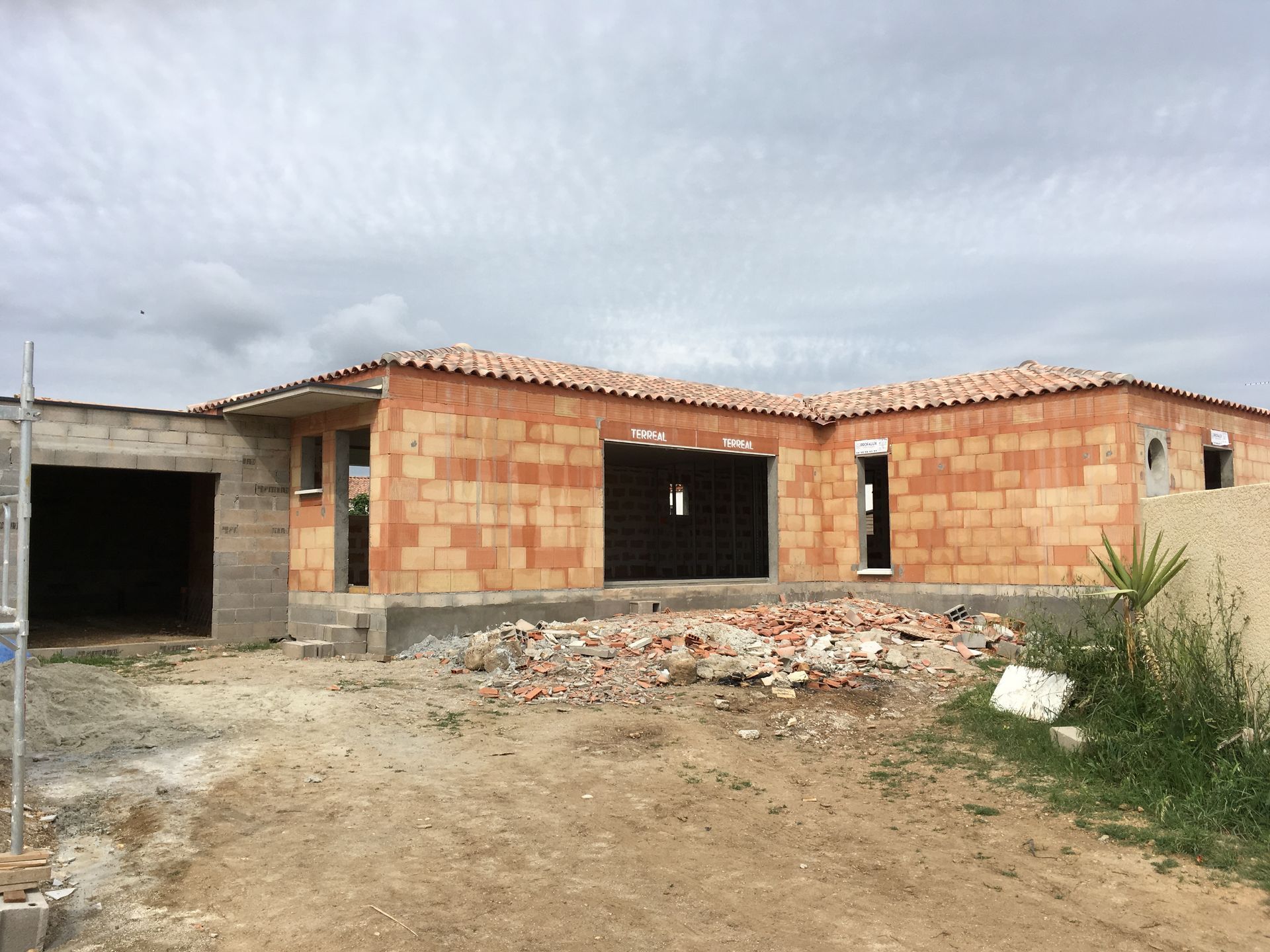 Maison en briques partiellement construite, avec toit inachevé et garage ouvert, sur un terrain en terre battue.