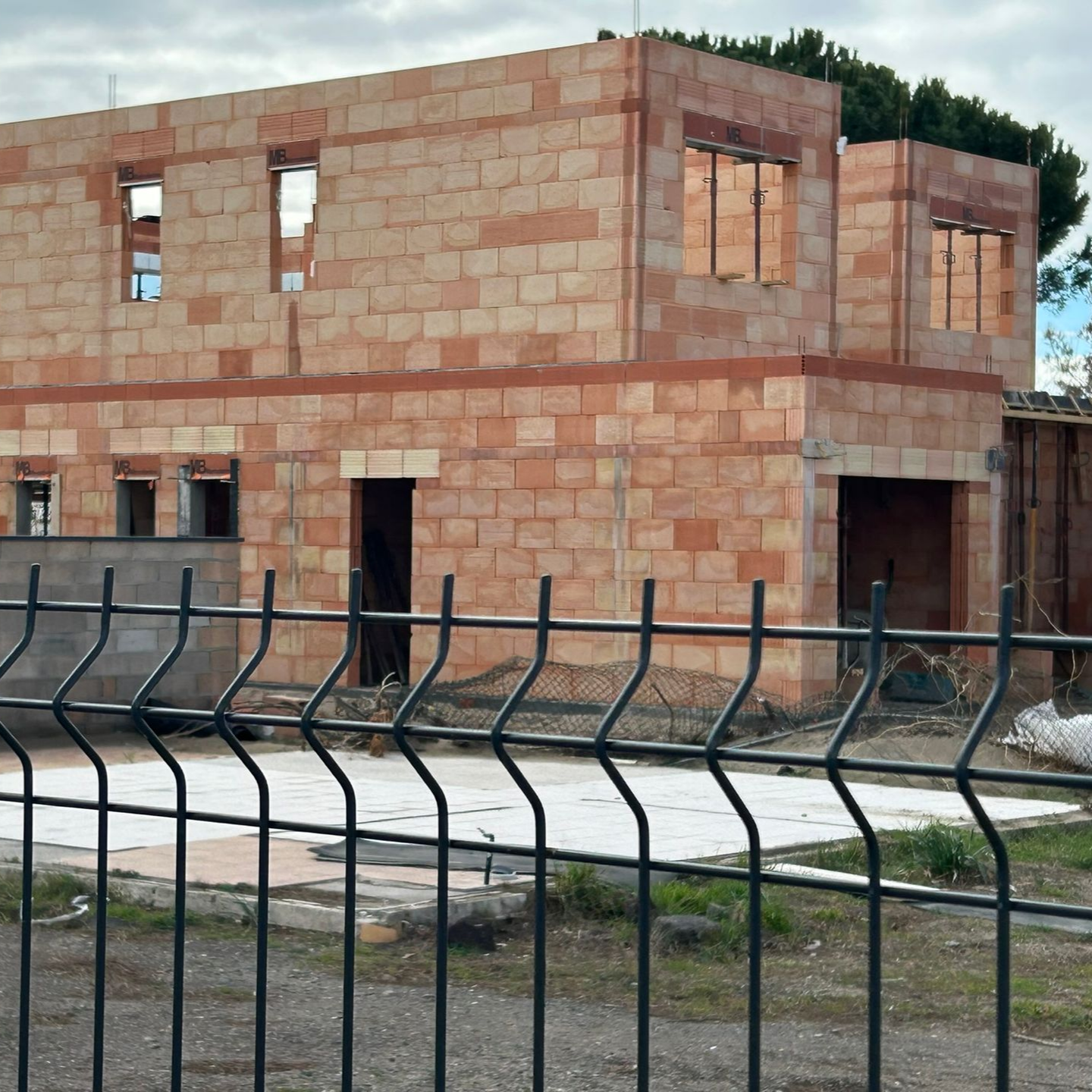 Maison en briques partiellement construite derrière une clôture en métal noir, avec des fenêtres et une toiture inachevées.