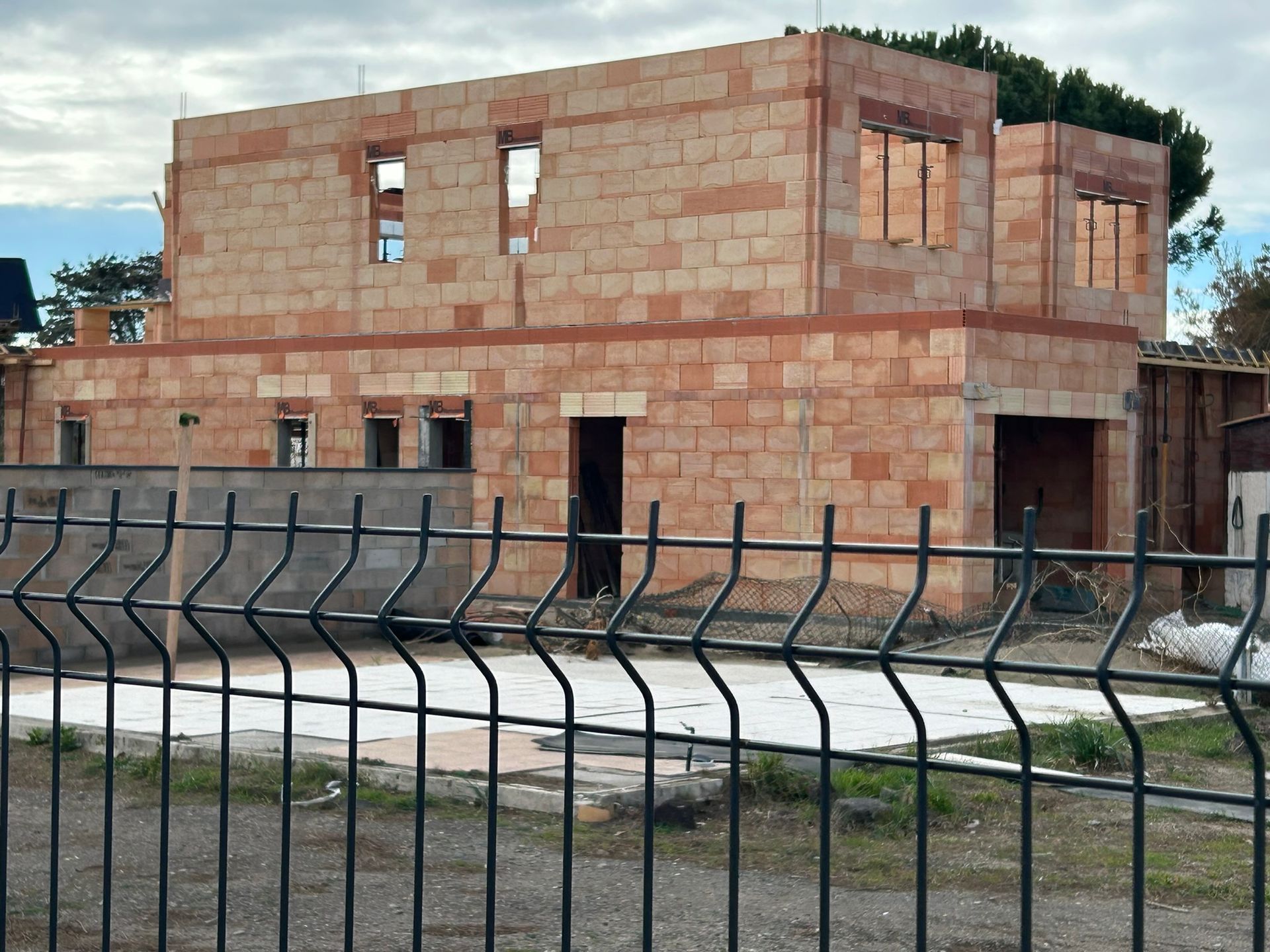 Maison en briques rouges partiellement construite derrière une clôture en métal noir sur un terrain gazonné