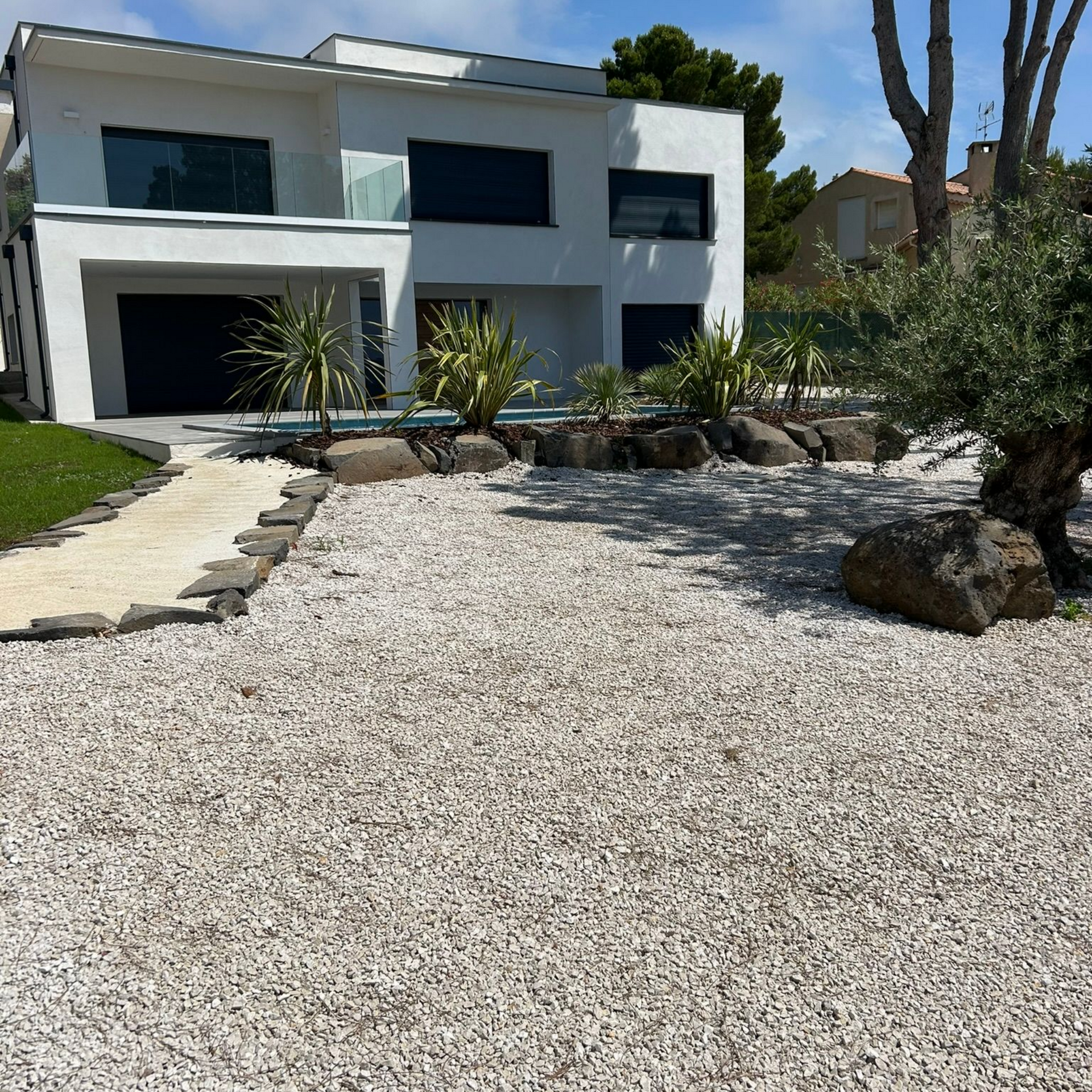 Maison moderne blanche de deux étages avec fenêtres sombres, cour avant en gravier et aménagement paysager désertique.