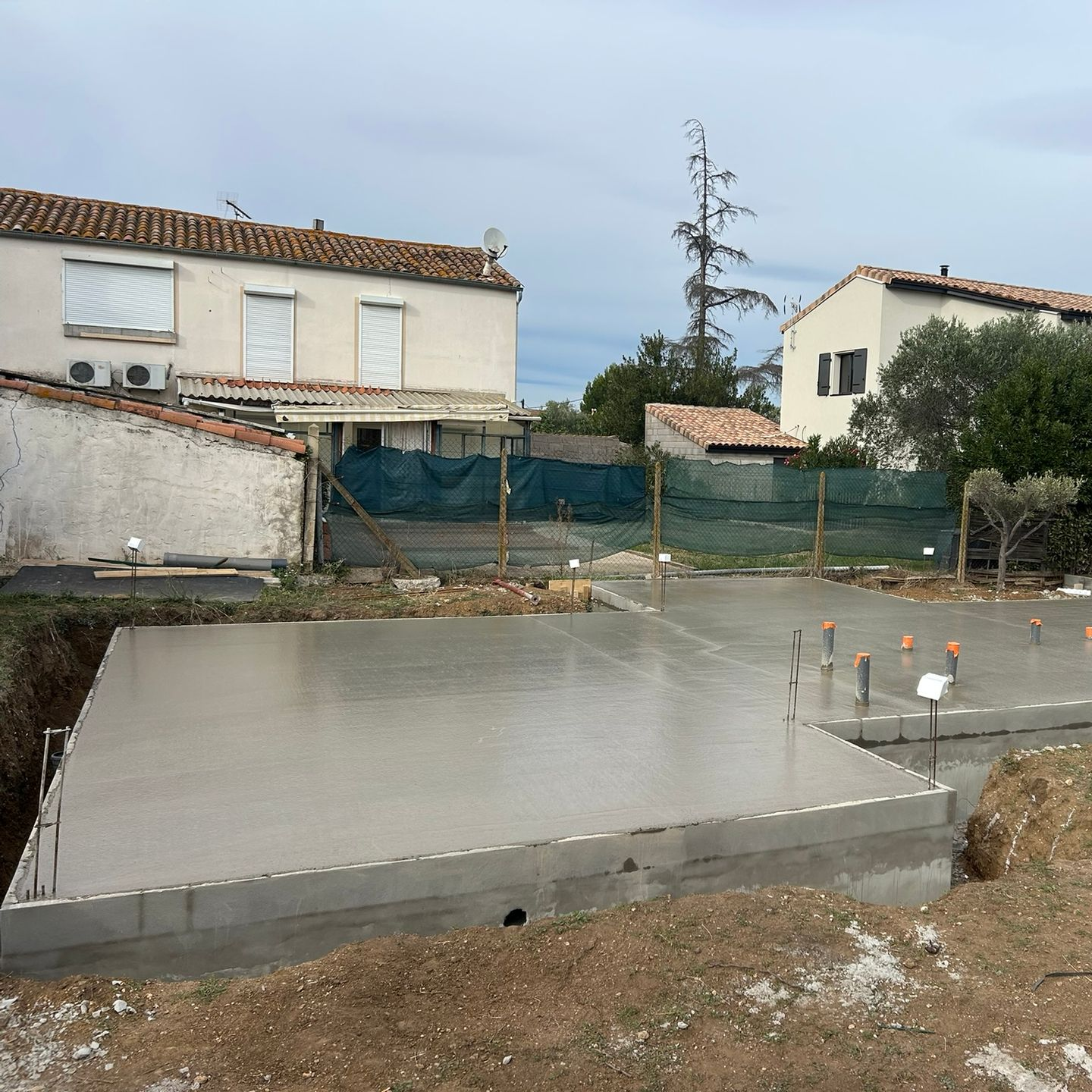 Dalle de fondations en béton en construction à côté de maisons, avec clôture et terrain boueux.