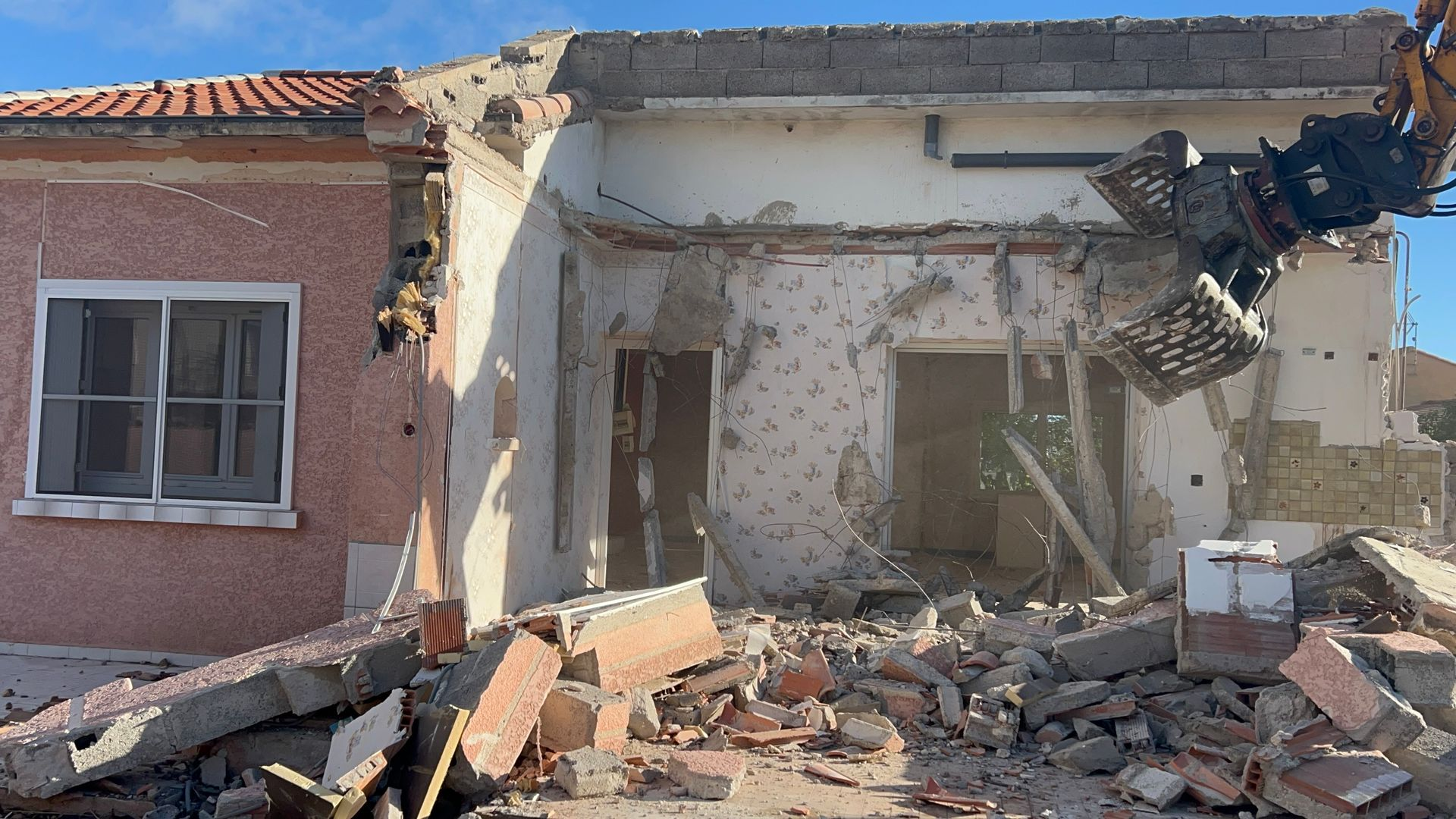 Maison partiellement démolie, avec des décombres, des murs intérieurs apparents et un bâtiment voisin rose sur la gauche.