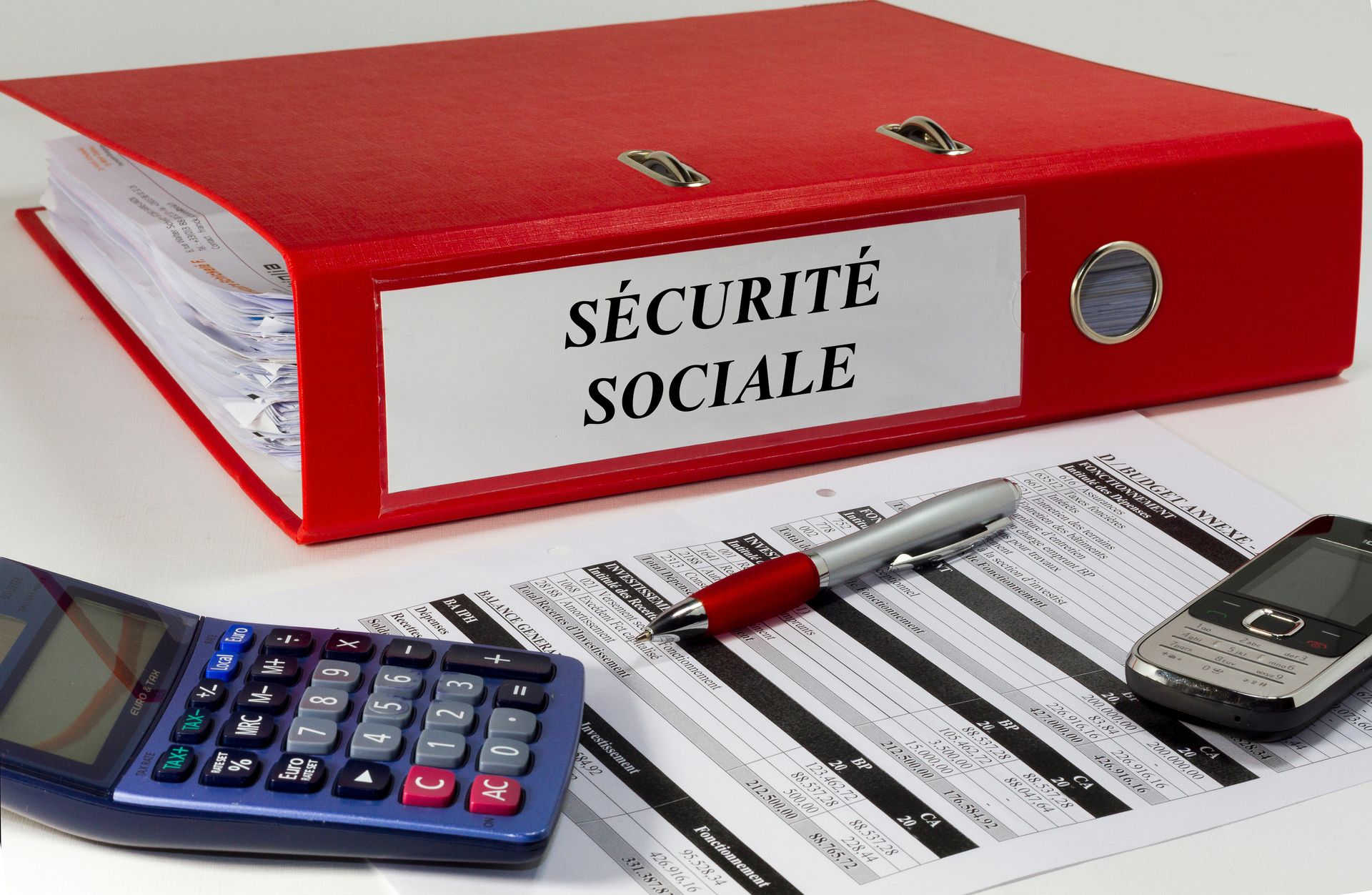 Dossier rouge Sécurité sociale contenant une calculatrice, un stylo, un document et une carte d’identité sur un bureau.