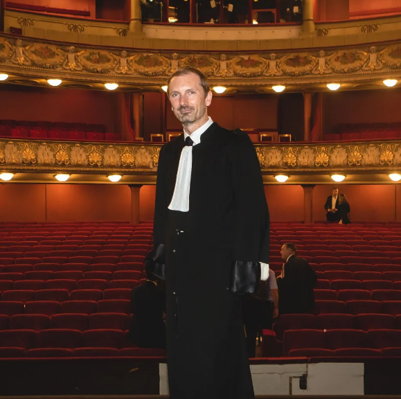Maître Vidal se tient debout dans un auditorium de théâtre, avec des sièges rouges.
