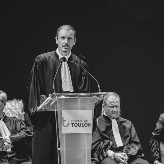 Maître Vidal en orateur à la tribune, micro en main, lors d'une cérémonie officielle.
