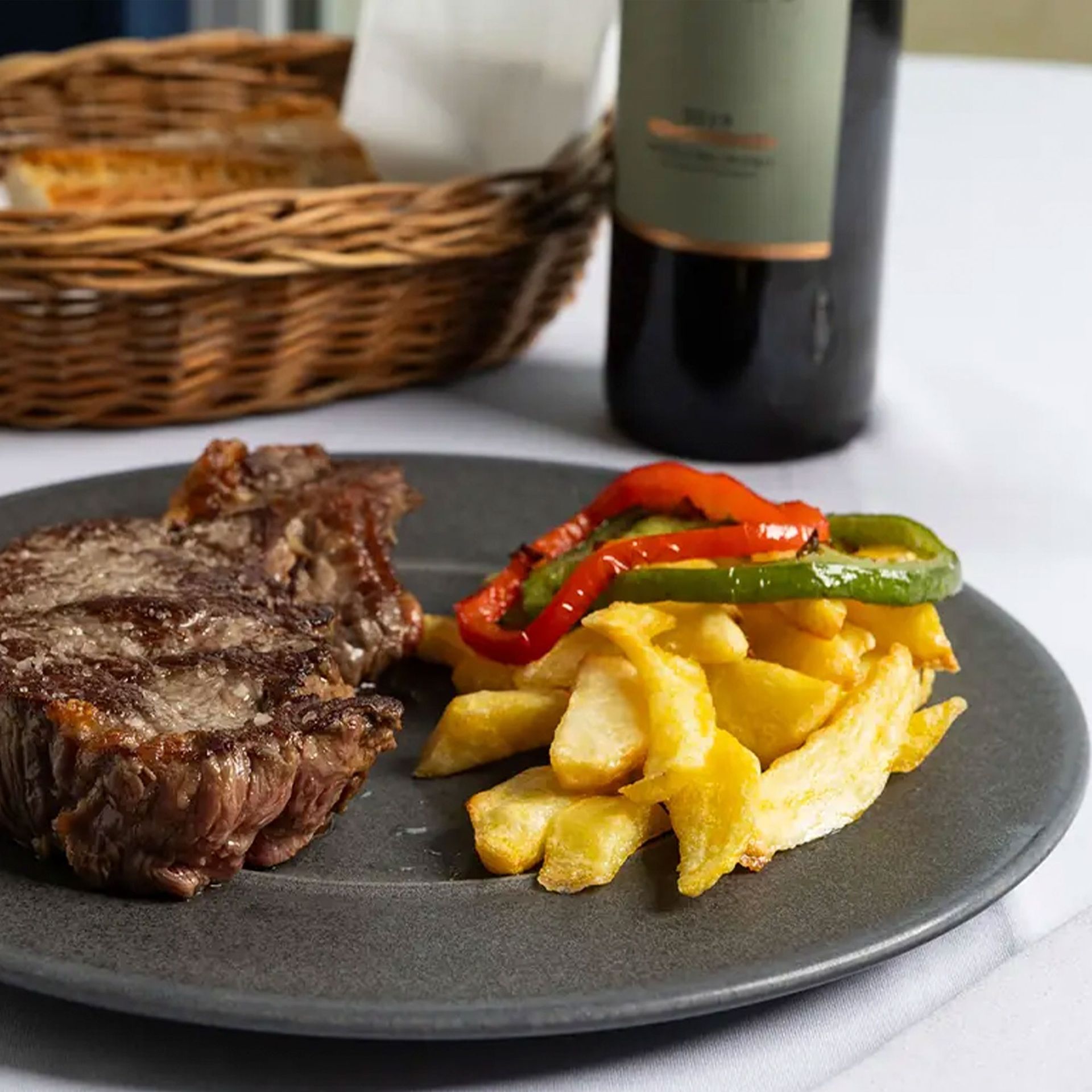 Filete con patatas asadas y pimientos rojos y verdes en un plato oscuro, con una cesta y una botella de vino detrás.