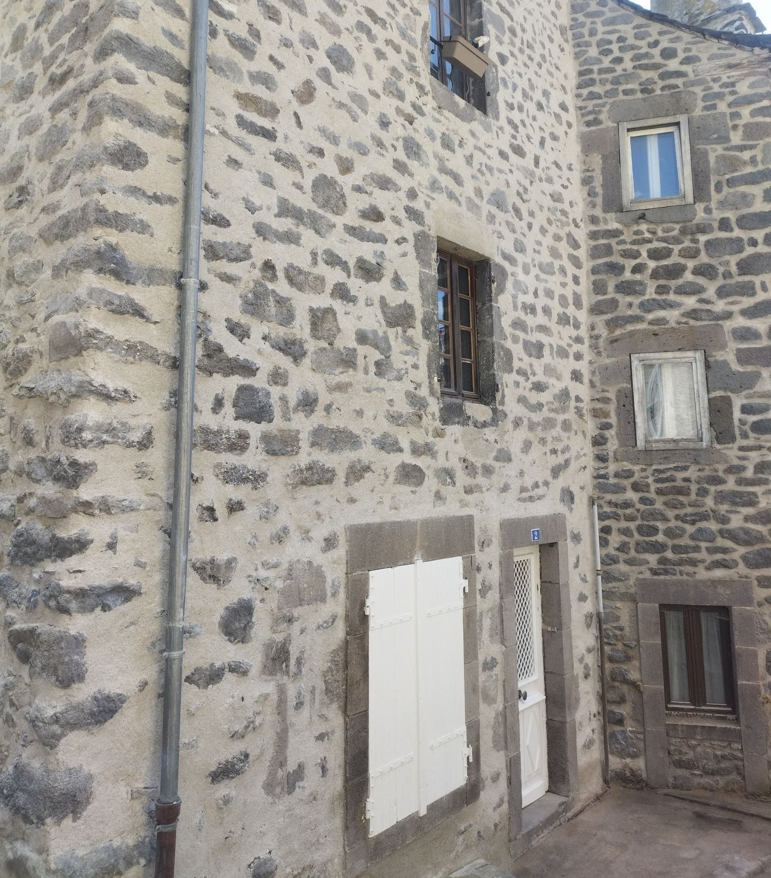 Façade en pierre avec volets blancs, fenêtres et gouttières métalliques.