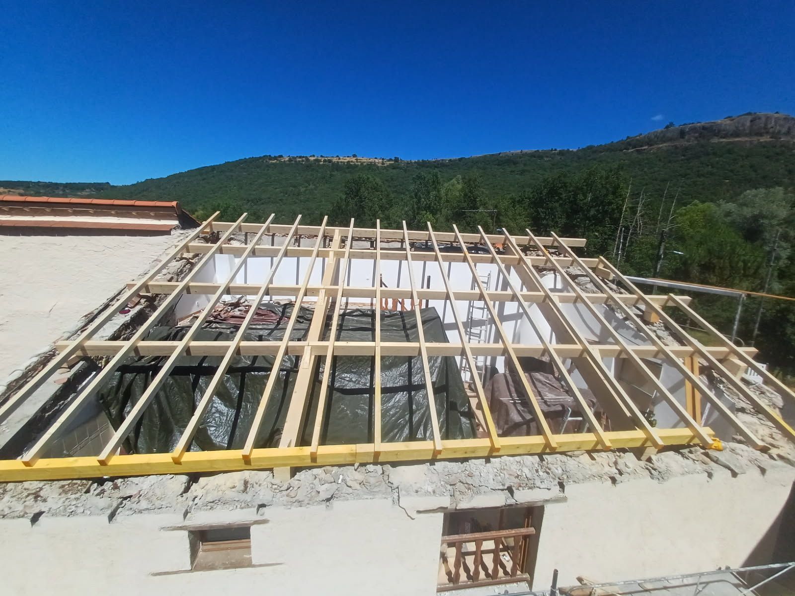 Toit d'un bâtiment en construction, charpente en bois, bâche verte, ciel bleu, montagnes en arrière-plan.