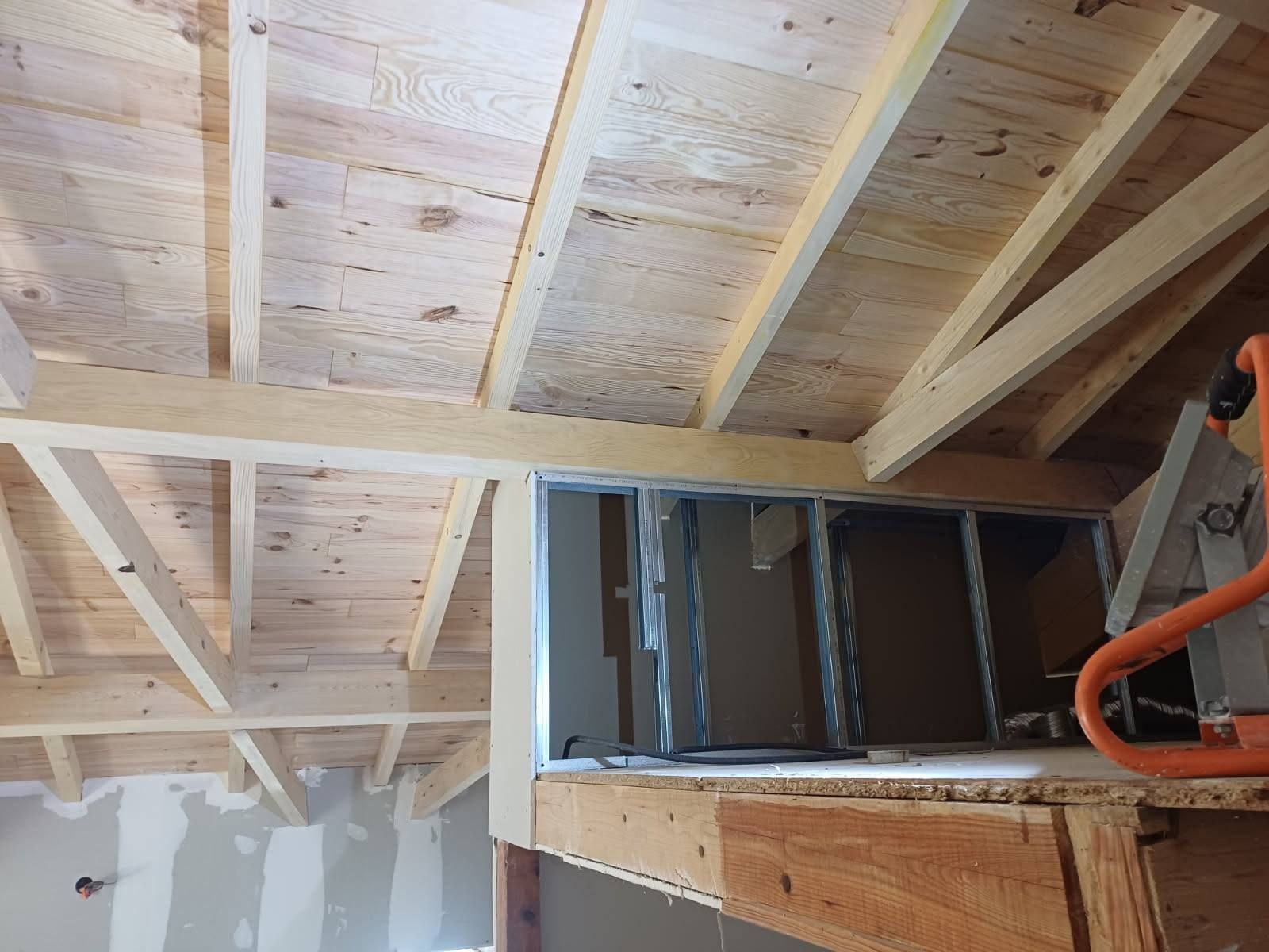 Charpente de plafond en bois avec une ouverture pour une fenêtre, vue d'en bas. Une scie circulaire repose sur un rebord.