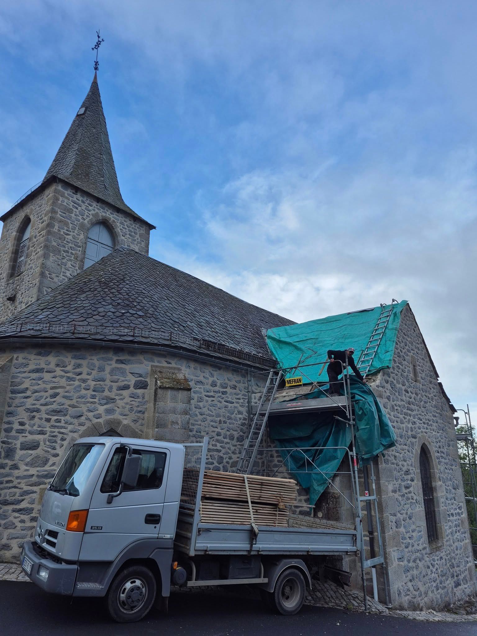 Un camion est stationné près d'une église en pierre dont le toit est en cours de réparation et recouvert d'une bâche verte, ciel nuageux.