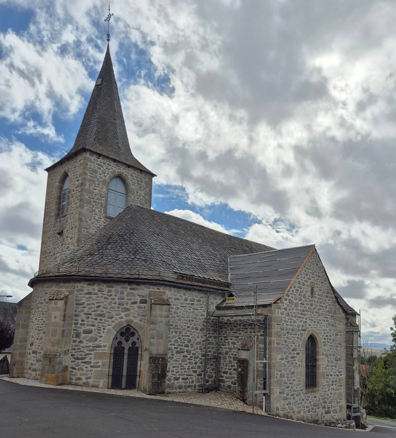 Église en pierre avec un haut clocher, des fenêtres cintrées et un toit d'ardoise sous un ciel nuageux.