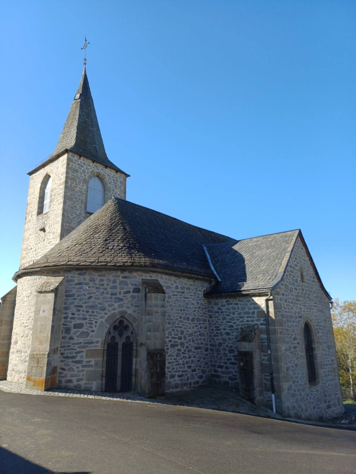 Église en pierre avec un haut clocher, un toit d'ardoise et des fenêtres cintrées se détachant sur un ciel bleu.