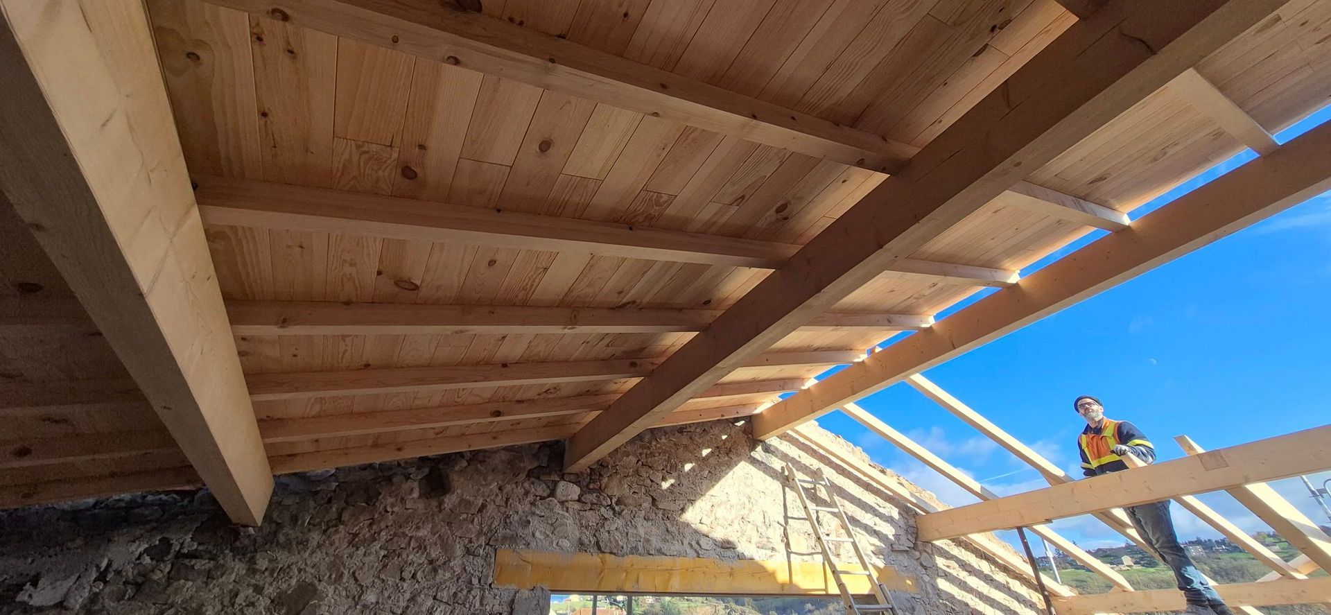 Un ouvrier du bâtiment travaille sur une charpente de toit en bois, avec un ciel dégagé en arrière-plan. Des planches sont en cours de pose.