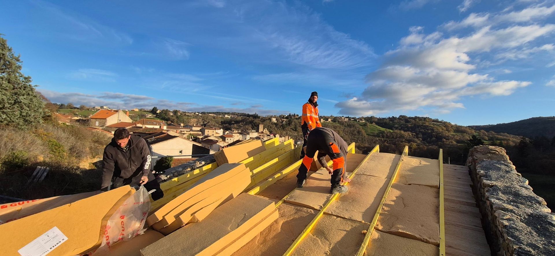 Des ouvriers installent de l'isolant sur un toit, avec vue sur une ville sous un ciel bleu.