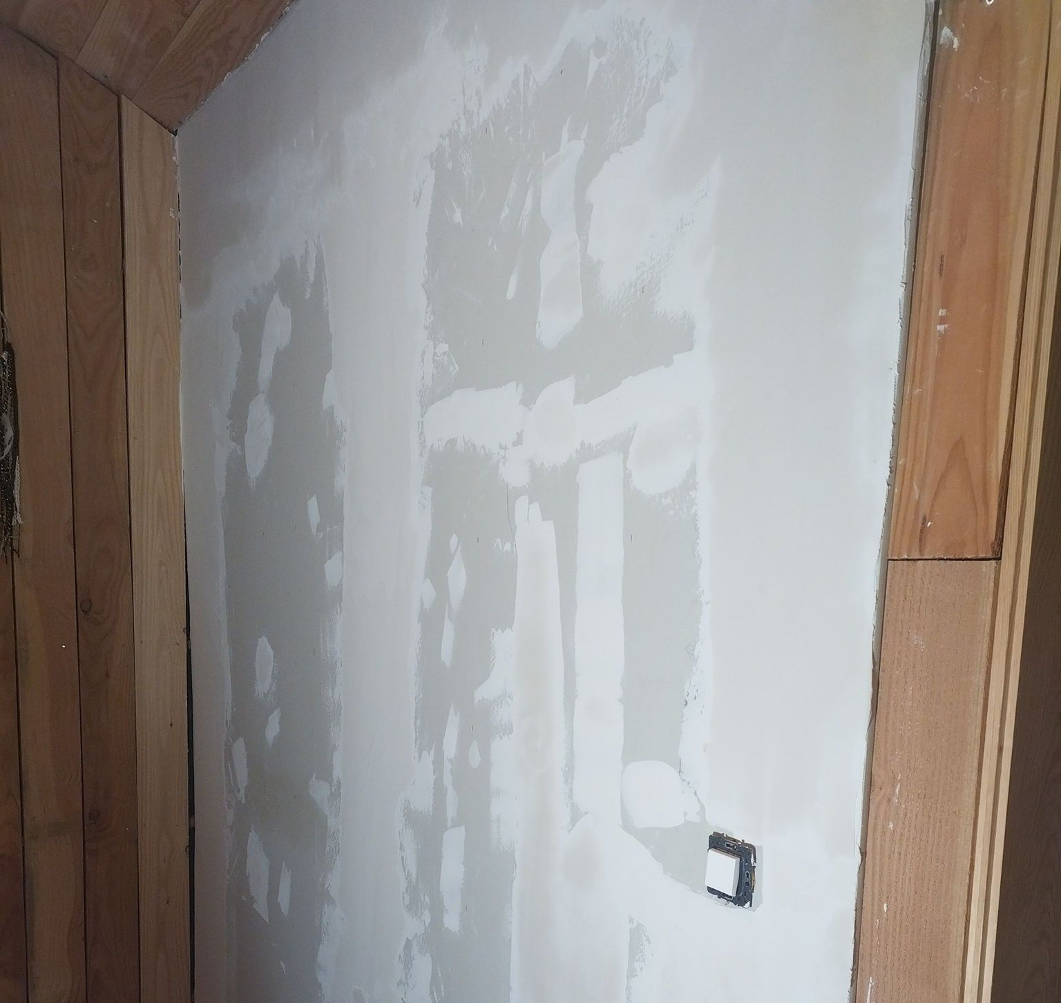 Mur réparé avec de l'enduit à plaques de plâtre, entouré de moulures en bois. Une prise électrique métallique est visible.