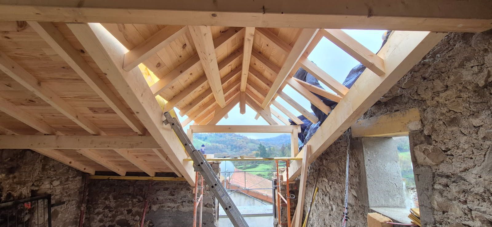 Vue intérieure d'un bâtiment en construction, avec des poutres en bois et un toit ouvert ; un ouvrier est visible.
