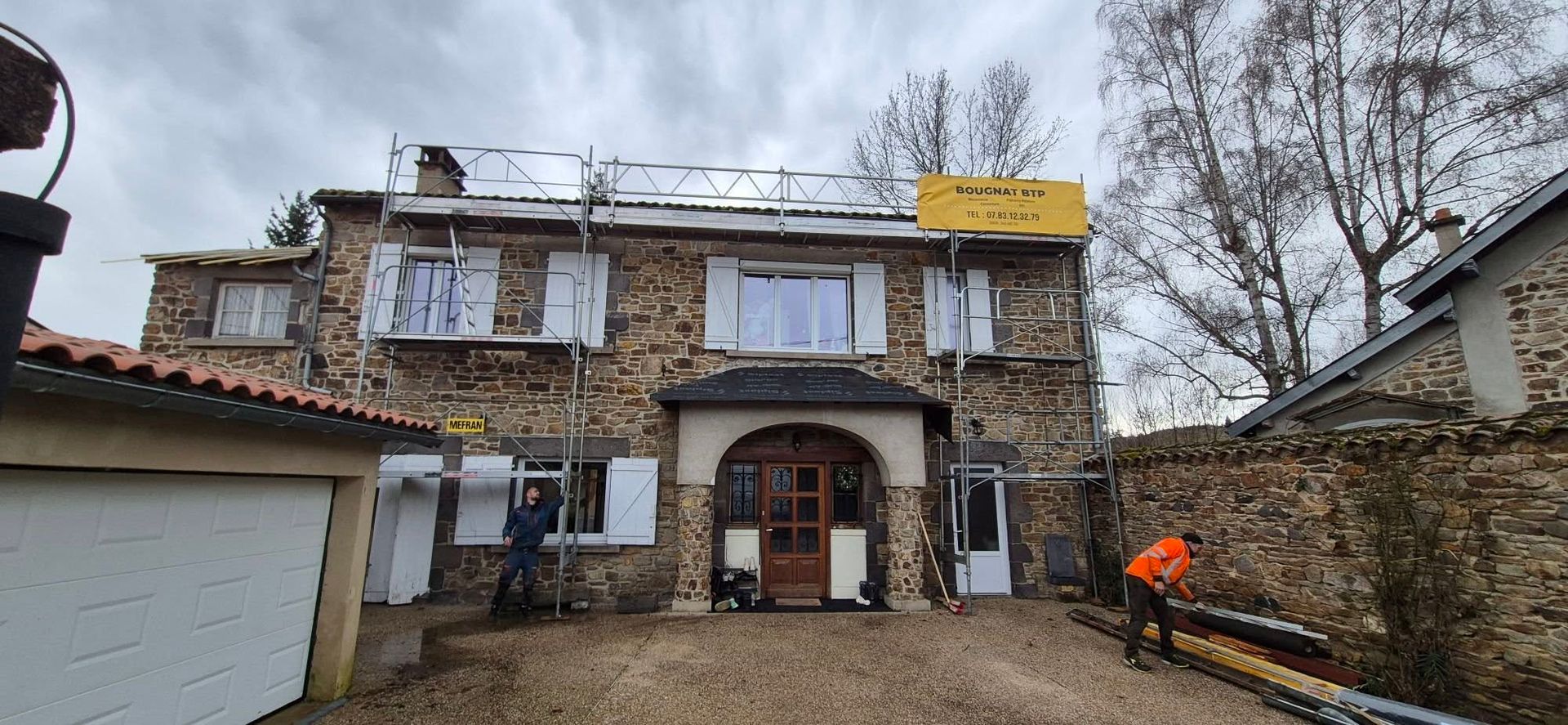Des ouvriers sur des échafaudages près d'une maison en pierre installent des panneaux solaires sous un ciel nuageux.