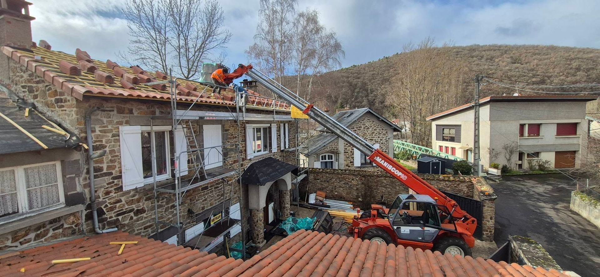 Des ouvriers utilisent une nacelle élévatrice pour réparer le toit d'un bâtiment en pierre ; un chariot élévateur orange se trouve devant le bâtiment.