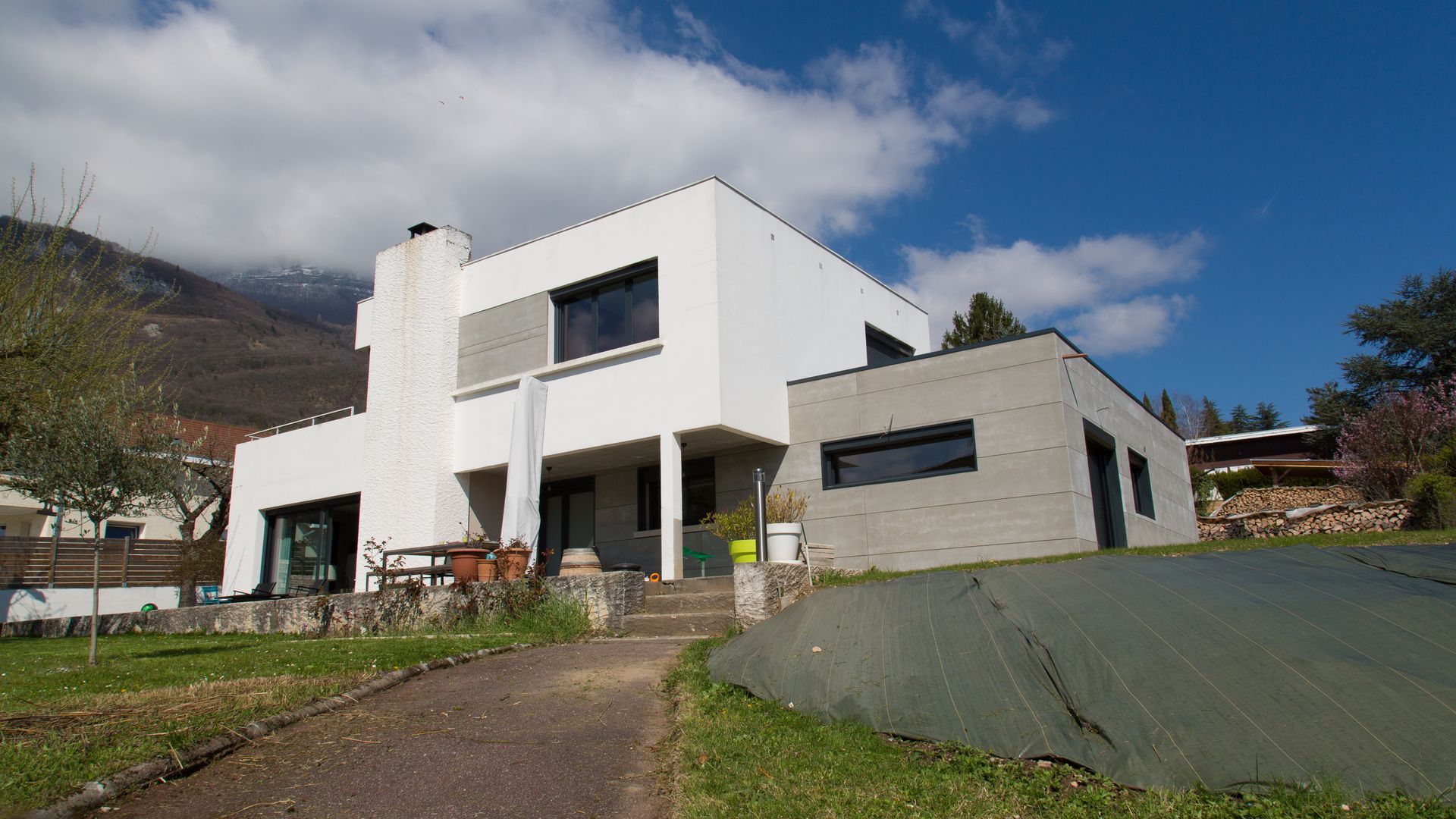Maison moderne blanche de deux étages avec toit plat, extension grise et jardin sous un ciel bleu.