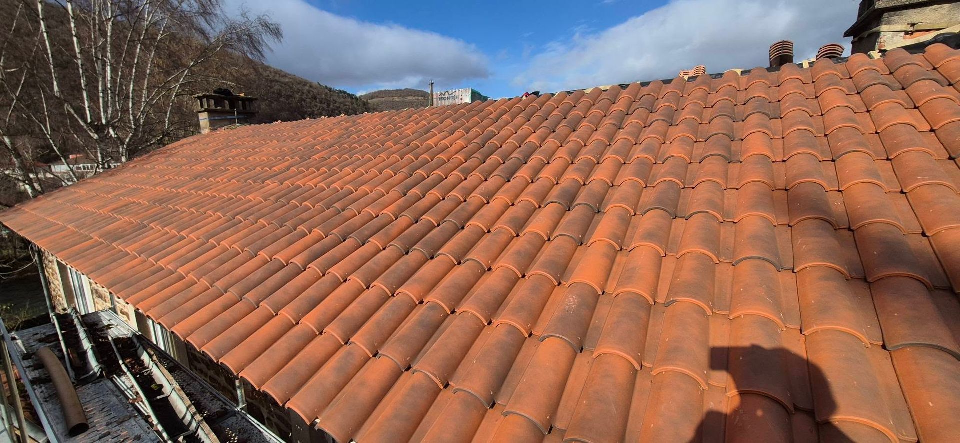 Toit de tuiles en terre cuite rouge, ciel bleu, arbres en arrière-plan.