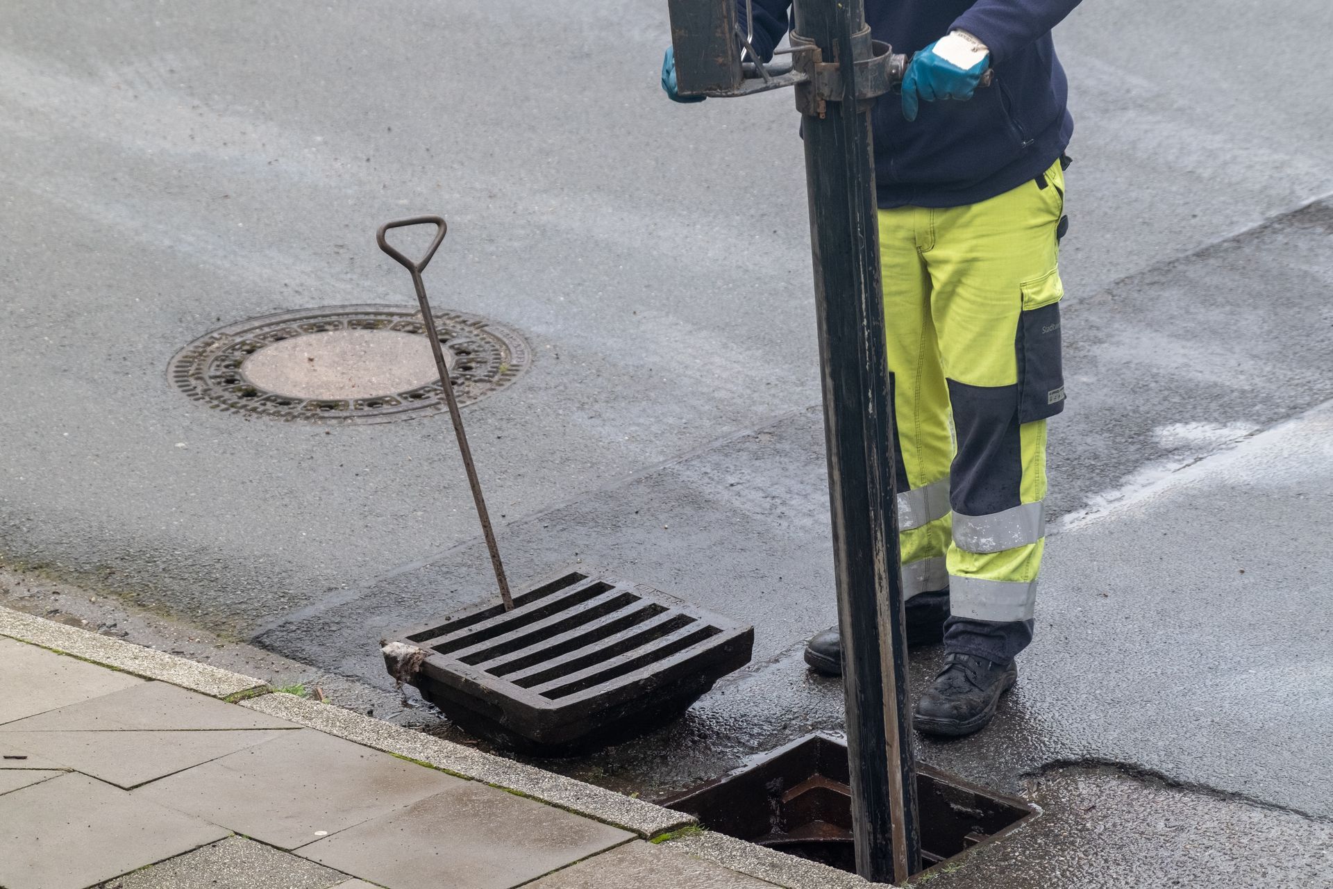 Eine Person in Schutzausrüstung öffnet einen Abwassergitter auf einer nassen Straße.